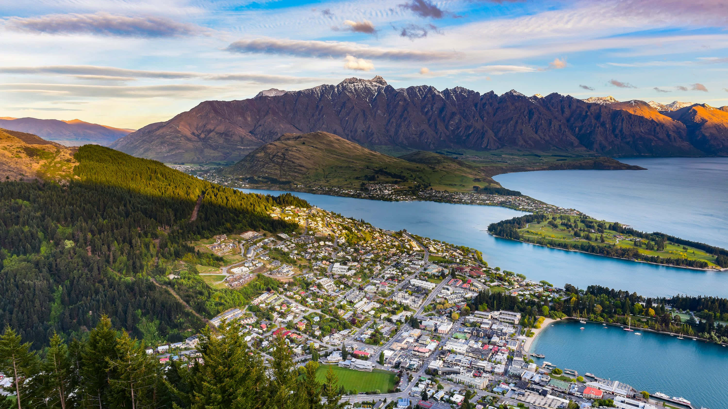 New Zealand 桌布 壁紙 — 從高空俯瞰，一座風景如畫的小鎮坐落在寧靜的藍色湖泊旁，背景中積雪覆蓋的山脈在黃金時刻雄偉地聳立。