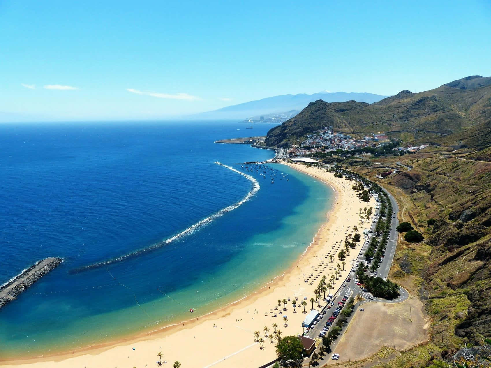 Spanish Beach 桌布 壁紙 — 從高空俯瞰，一條充滿活力的綠松石色海灘沿著崎嶇的海岸線蜿蜒，小鎮依偎在丘陵之中，天空湛藍清澈。
