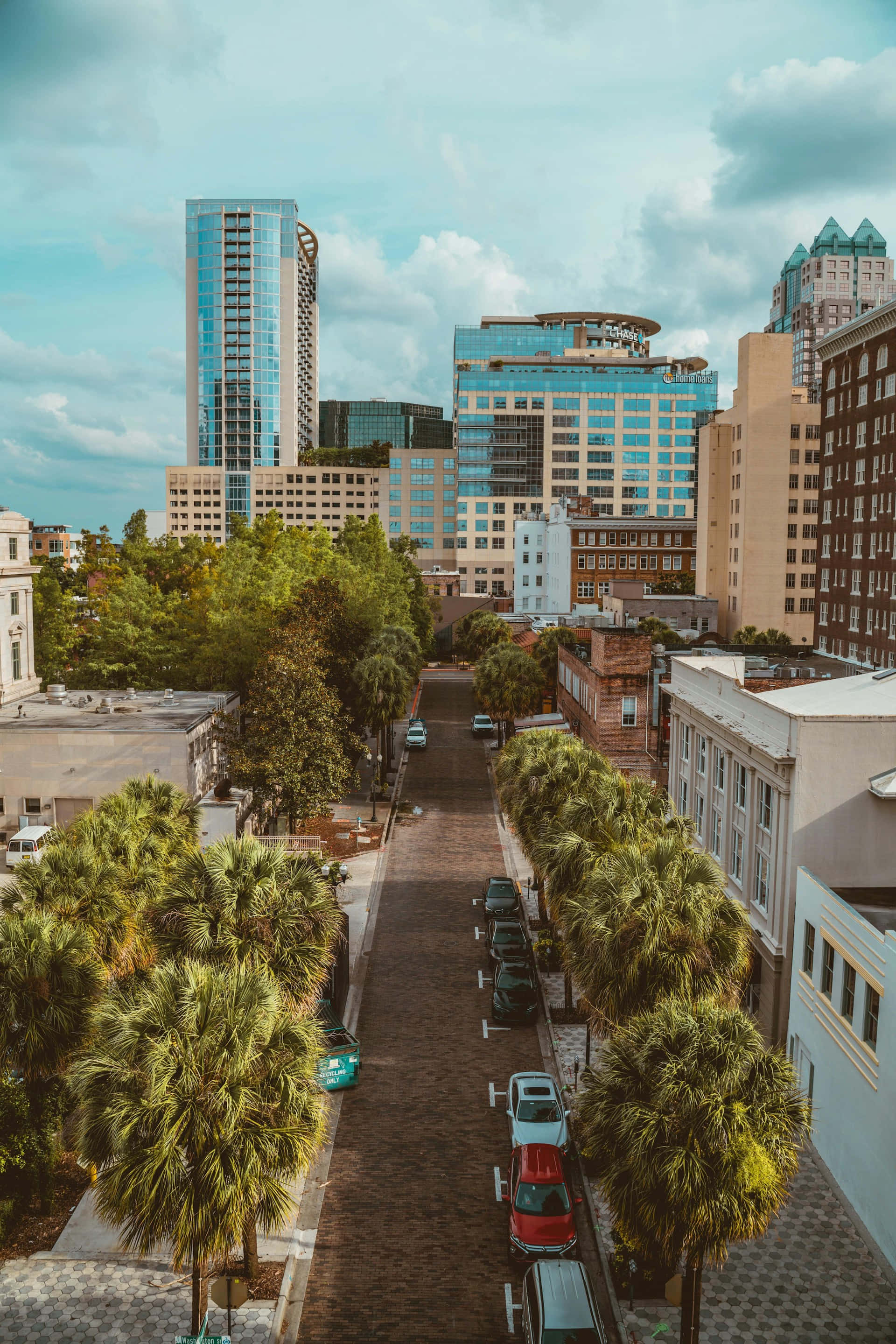 Orlando 桌布 壁紙 — 從高空俯瞰，一條綠樹成蔭的磚砌街道蜿蜒穿過充滿活力的城市景觀，現代玻璃摩天大樓和歷史建築在部分多雲的天空下交相輝映。