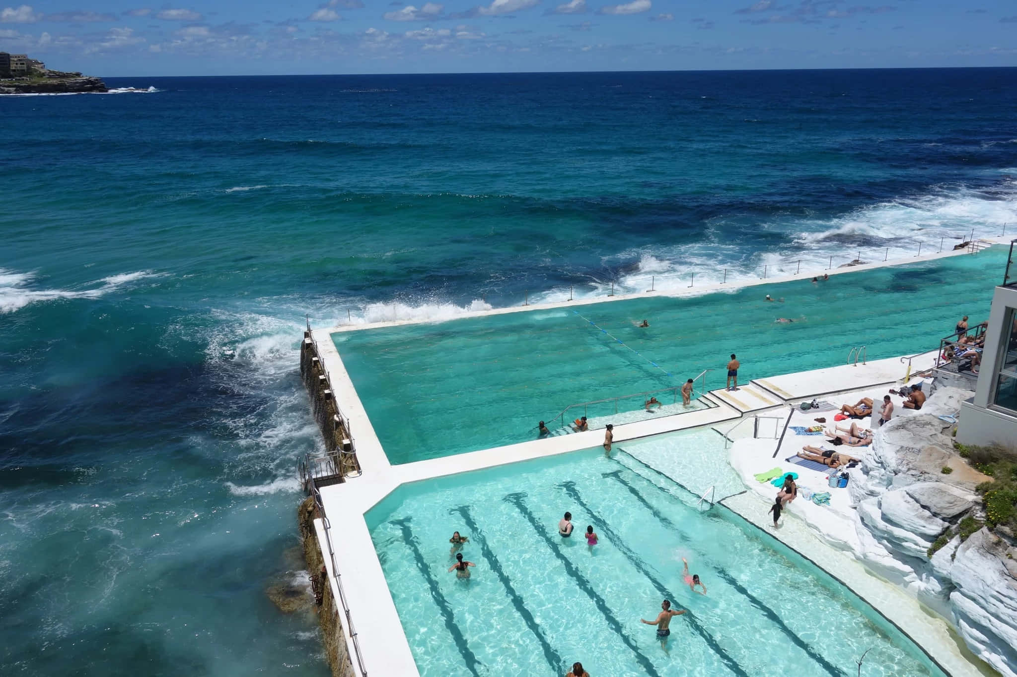 Beach Swimming Pool 桌布 壁紙 — 從高空俯瞰，在清澈藍天下，洶湧海浪旁有一個充滿活力的藍綠色海洋泳池，裡面有游泳者和日光浴者。