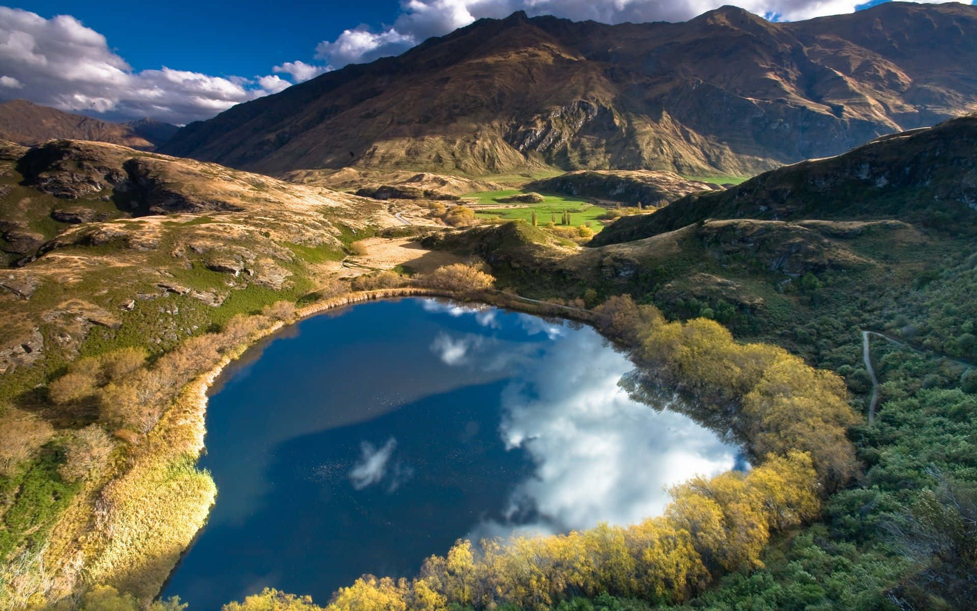 New Zealand 桌布 壁紙 — 從高空俯瞰，寧靜的山間湖泊在明亮的藍天下倒映著雲朵，周圍環繞著秋日樹木和崎嶇地形。