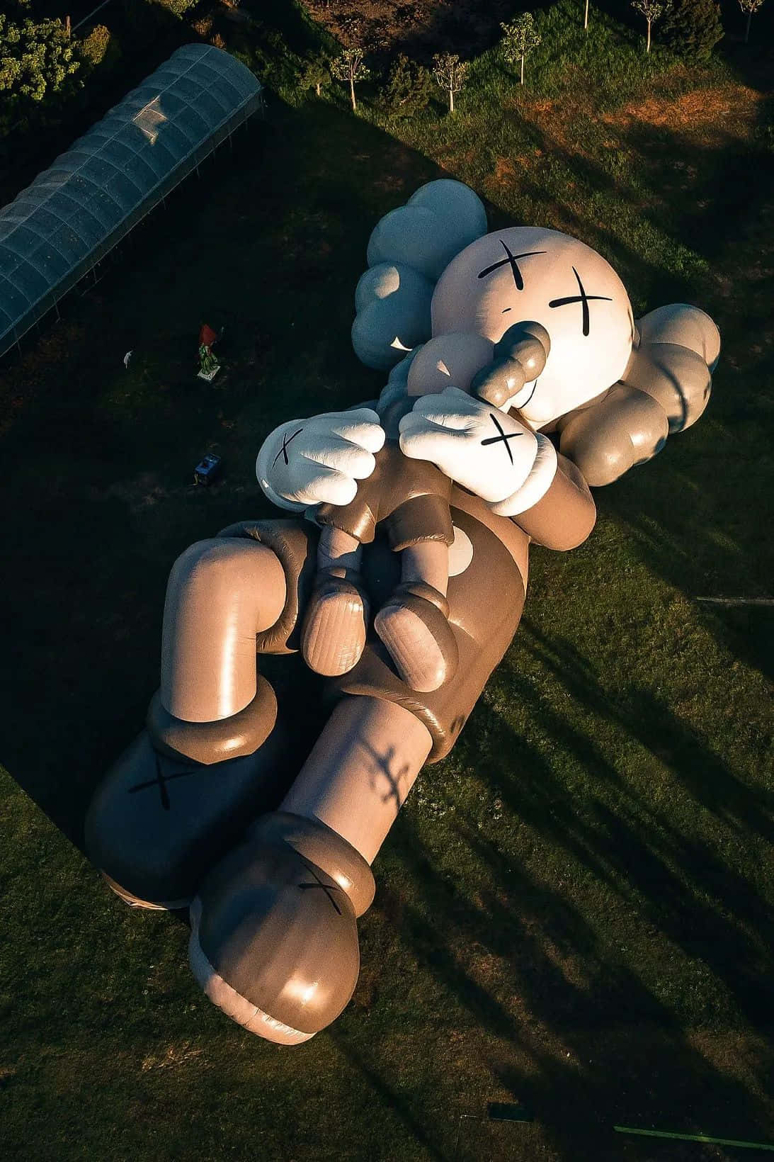 Kaws Holiday 桌布 壁紙 — 從高空俯瞰，巨大的KAWS Companion雕塑躺在草地上，長長的影子延伸穿過田野。