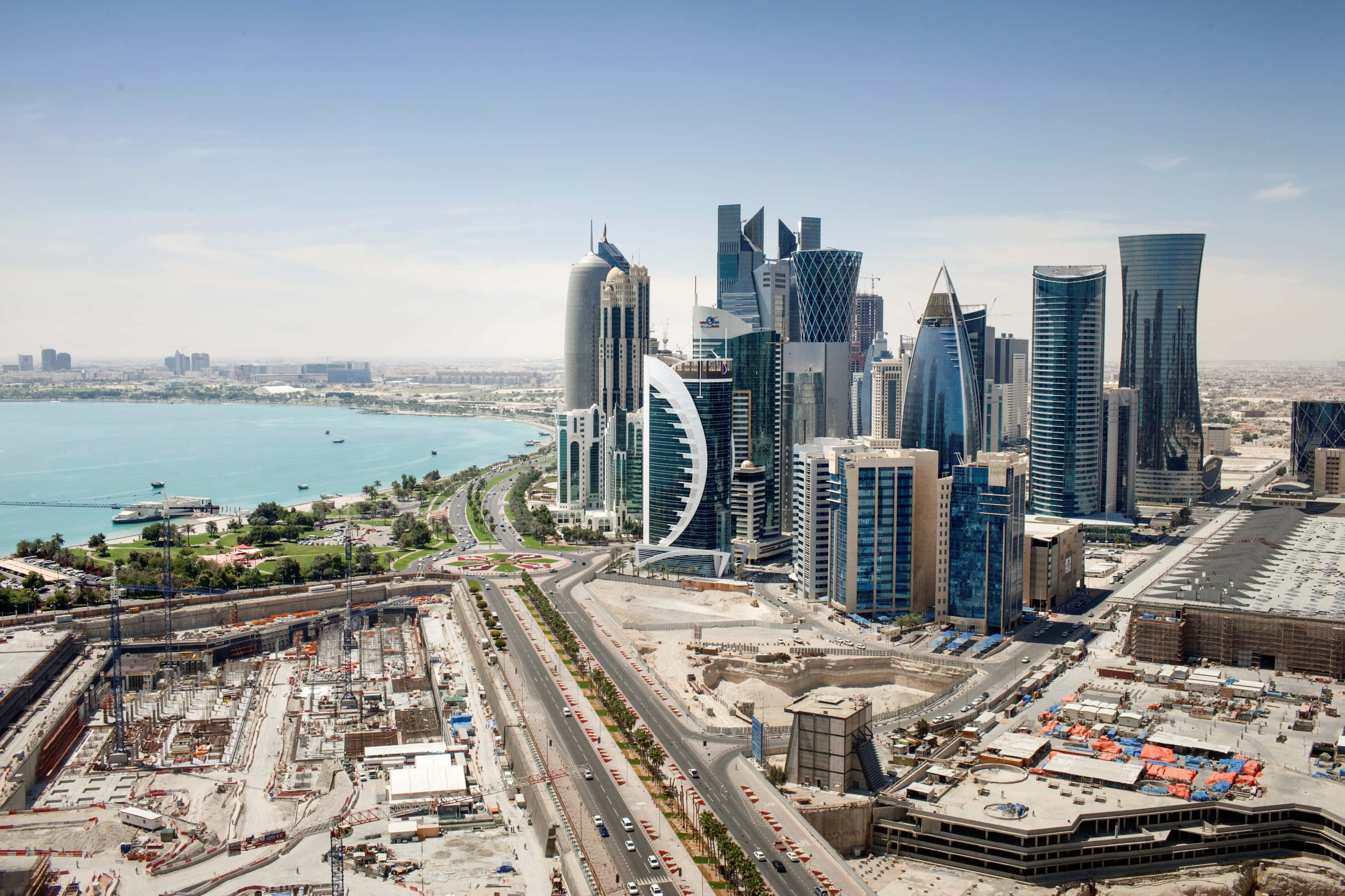 Qatar 桌布 壁紙 — 從高空俯瞰，未來主義摩天大樓沿著碧綠海灣排列，下方有活躍的建築工地和道路，在晴朗藍天下呈現。