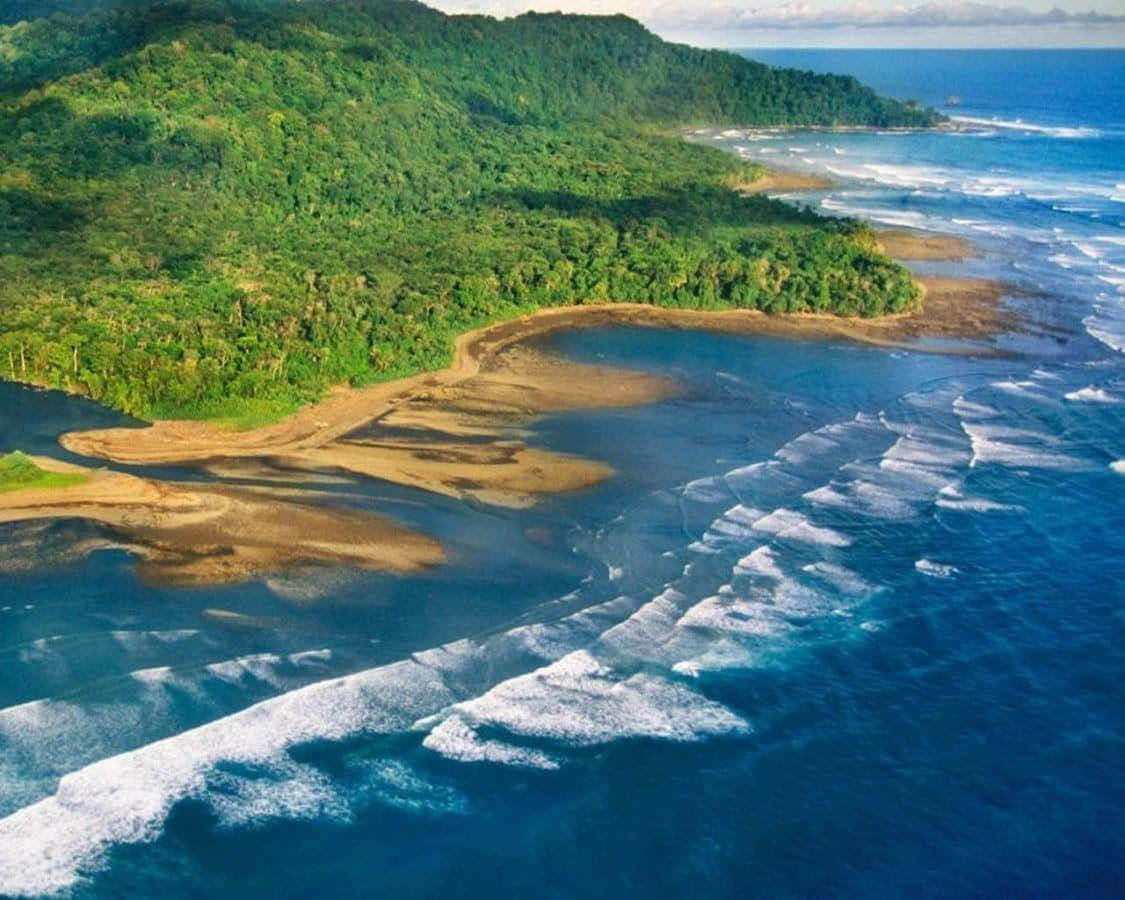 Costa Rica 桌布 壁紙 — 從高空俯瞰，碧綠色的海浪拍打著一個沙灣，這個沙灣坐落在茂密的綠色叢林山丘和深藍色的海洋之間。