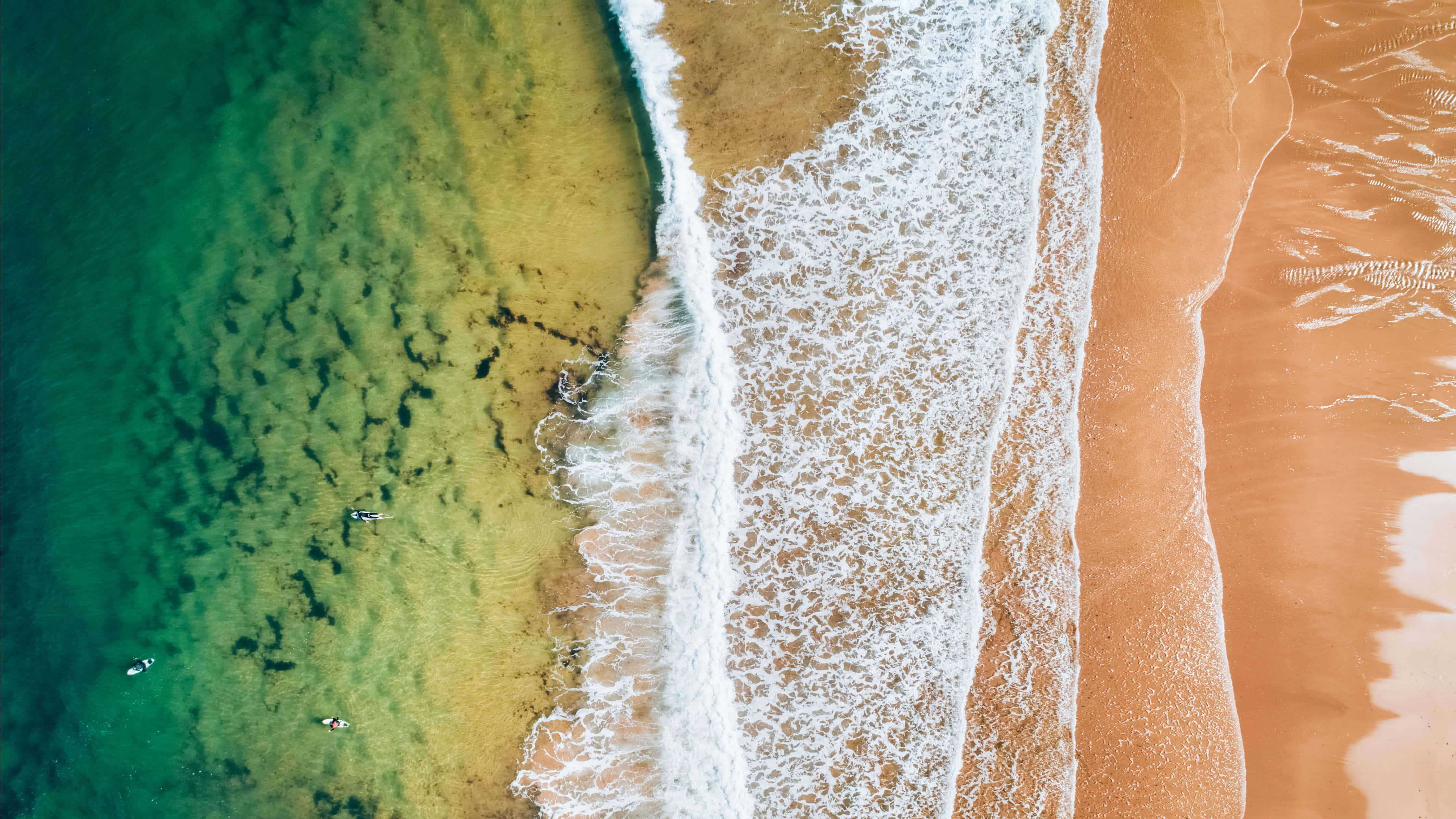 Beach Photography 桌布 壁紙 — 從高空俯瞰，綠松石色的海浪拍打著金色沙灘，衝浪者在淺水中漂浮。