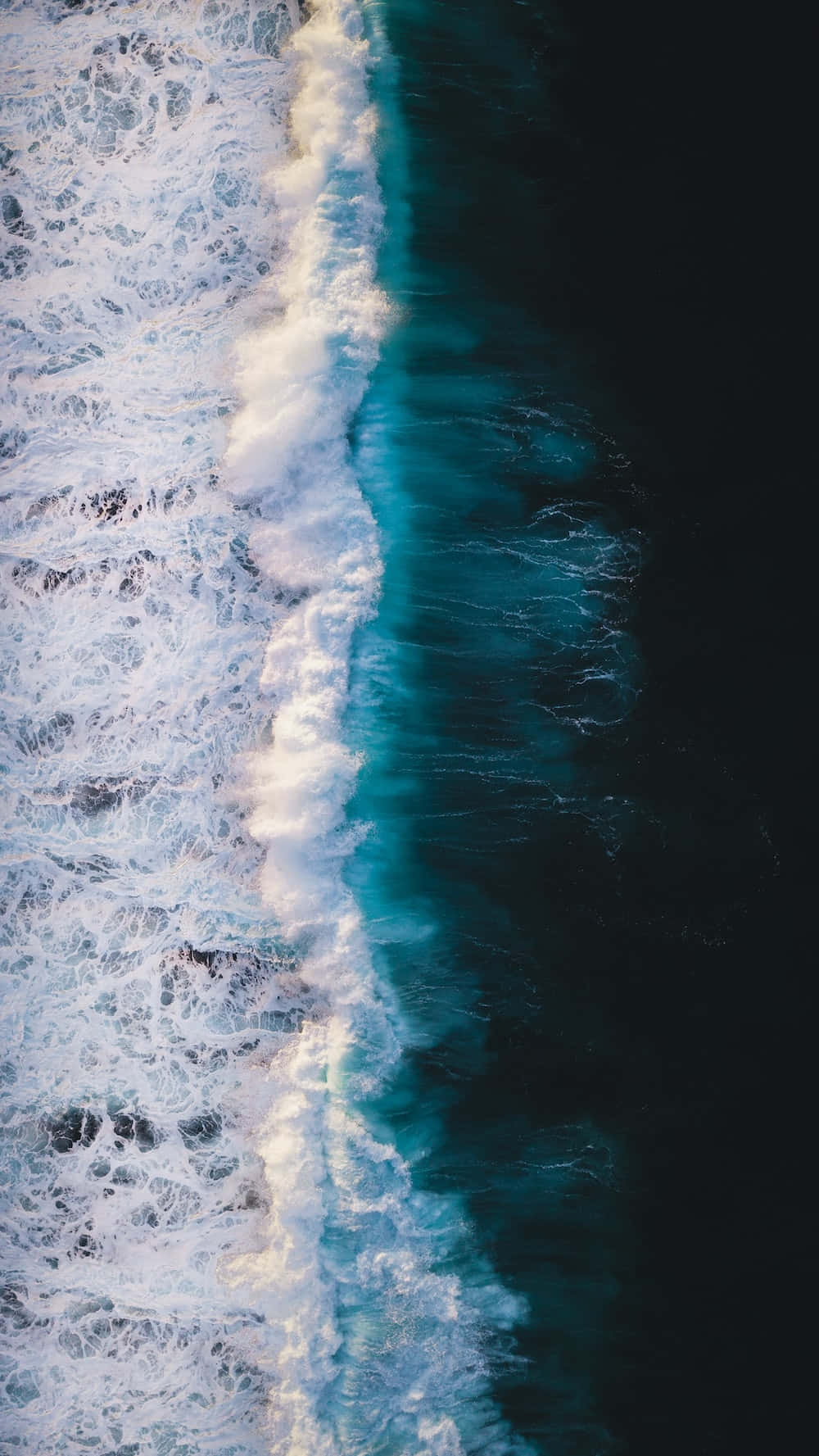 Portrait Mode 桌布 壁紙 — 從高空俯瞰，綠松石色的海浪拍打著深色海水，形成戲劇性的海岸線對比。