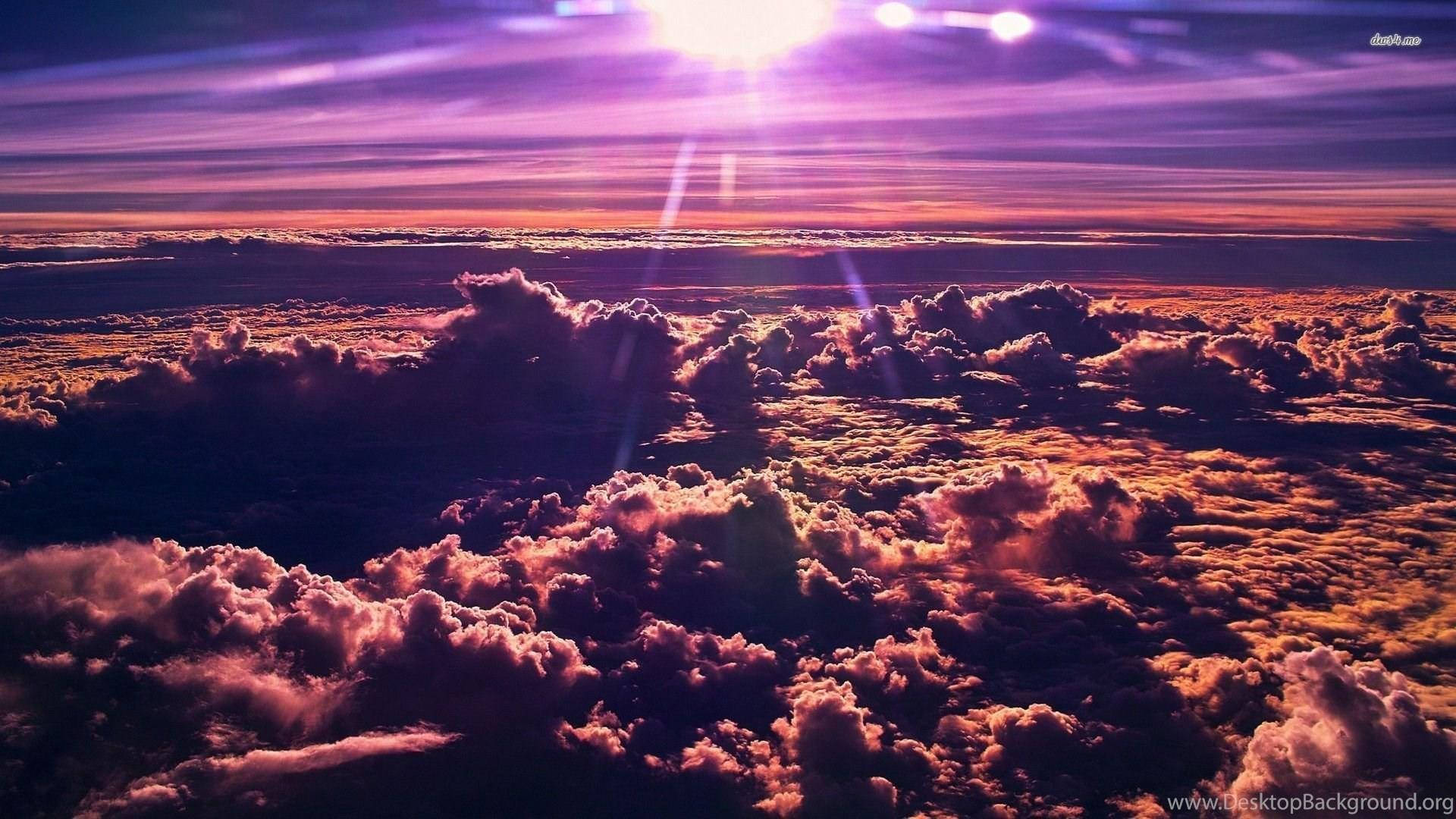 雲朵 桌布 壁紙 — 從高空俯瞰，蓬鬆的雲朵沐浴在充滿活力的紫色和橙色日落光芒中，燦爛的陽光穿透雲層。