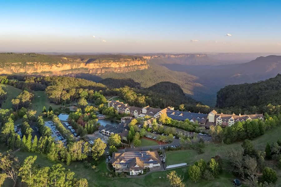 Blue Mountains National Park 桌布 壁紙 — 從高空俯瞰一個度假村，坐落於鬱鬱蔥蔥的山谷中，懸崖和遠山沐浴在金色的日落光芒中。