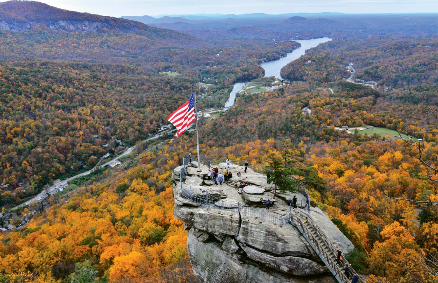 Fall Hikes 桌布 壁紙 — 從高空俯瞰岩石觀景點，美國國旗在秋日森林和蜿蜒河流上方飄揚。