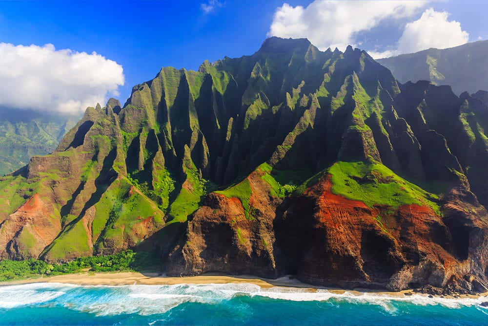 Hawaiian Island 桌布 壁紙 — 從高空俯瞰戲劇性的綠色和紅色懸崖，在明亮的藍天和蓬鬆的雲朵下，墜入碧綠色的海浪中。