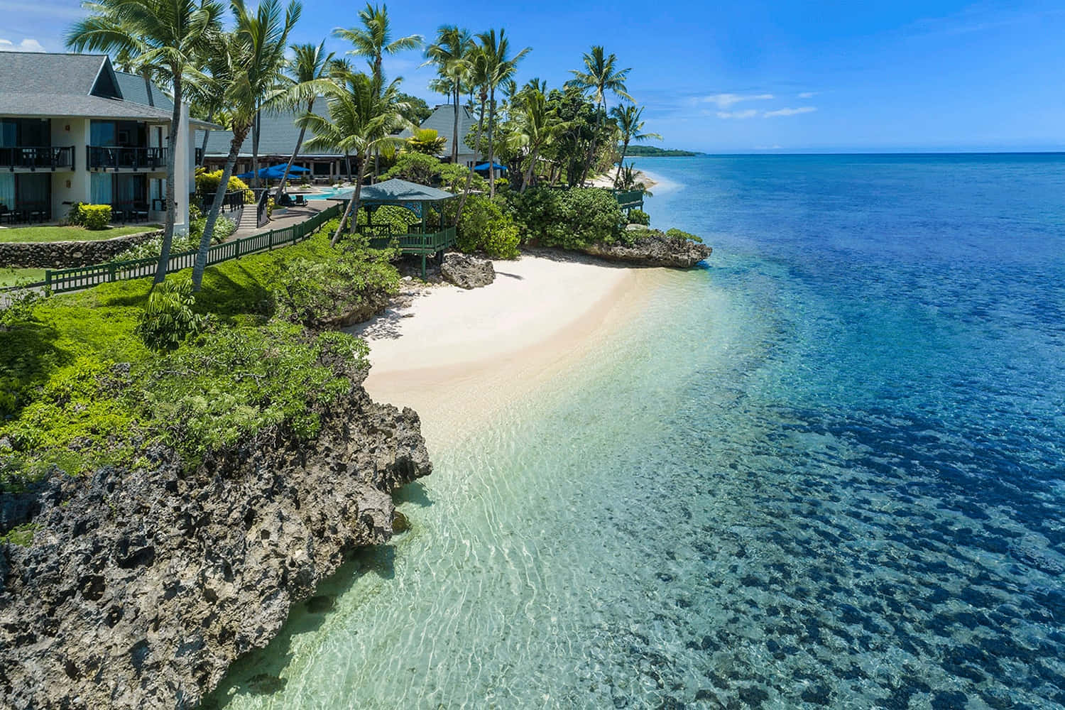 Fiji Beach 桌布 壁紙 — 從高空俯瞰熱帶海灘，白色沙灘、碧綠海水和棕櫚樹環繞度假村，天空湛藍明亮。
