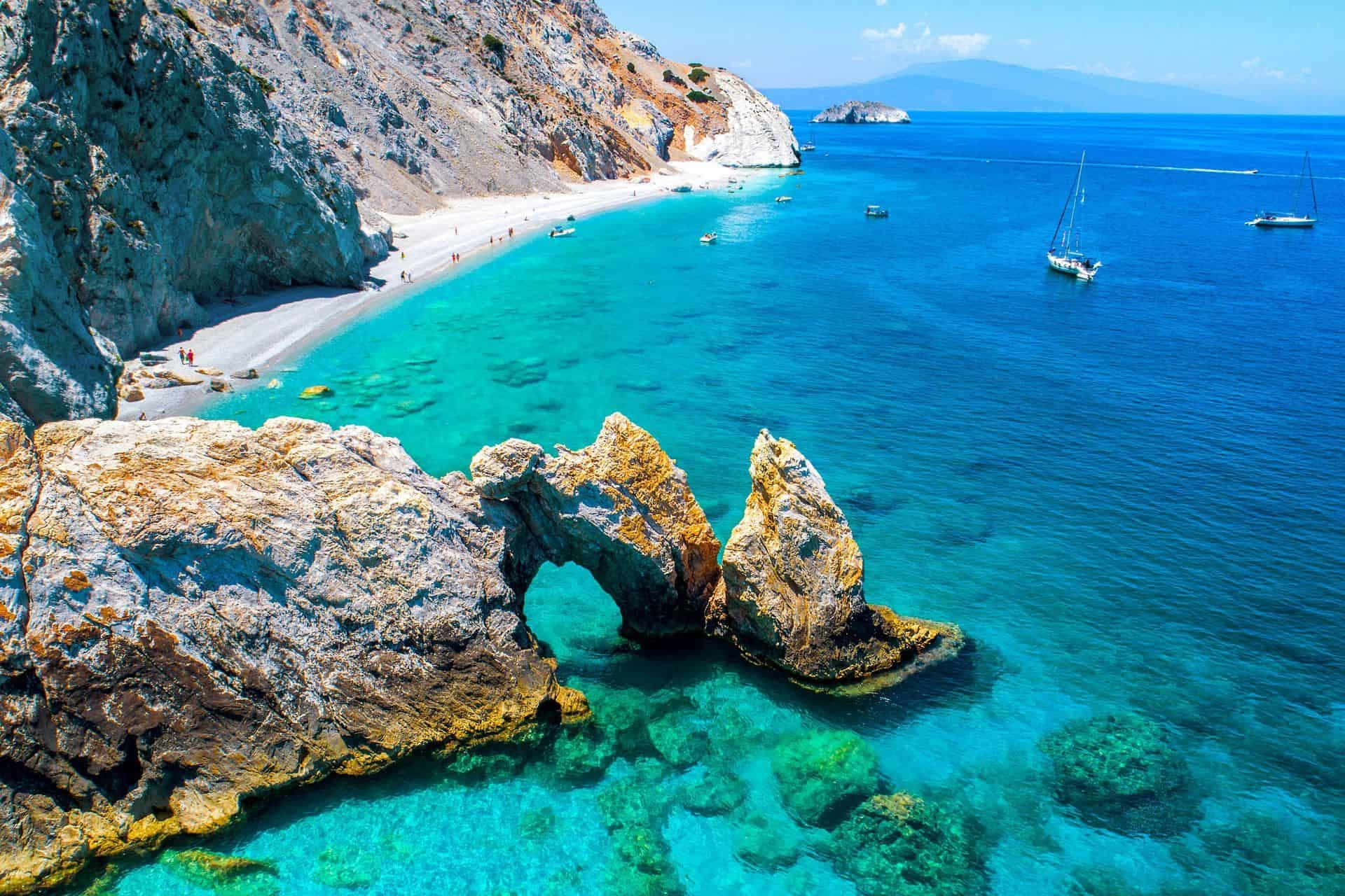 Greek Beach 桌布 壁紙 — 從高空俯瞰迷人的藍綠色海灣，天然岩石拱門和帆船點綴在充滿活力的藍色海洋上。