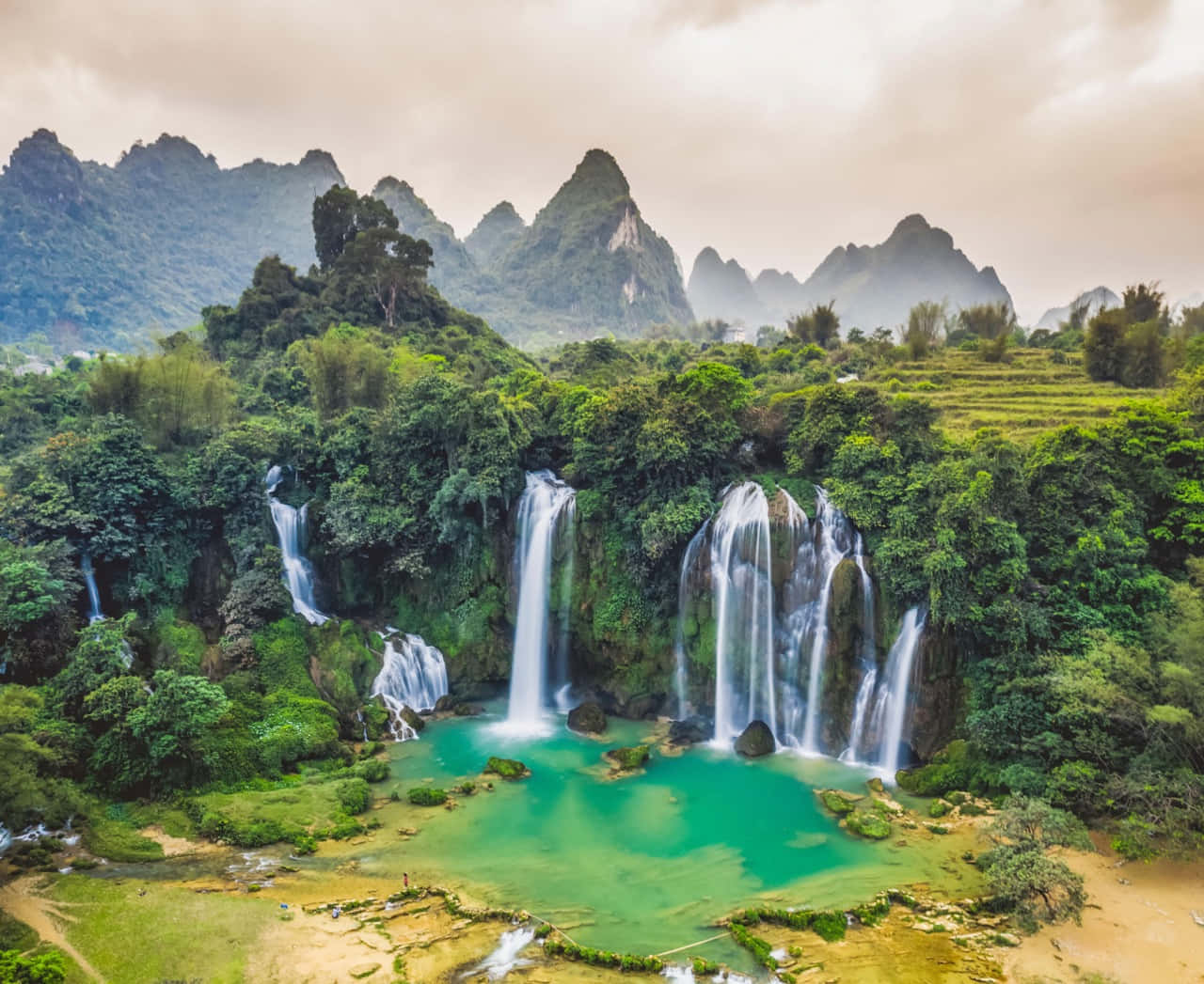 Eco-tourism 桌布 壁紙 — 從高空俯瞰雄偉的瀑布傾瀉而下，流入充滿活力的碧綠色水池，周圍環繞著茂密的綠色叢林和霧氣繚繞的喀斯特山脈。