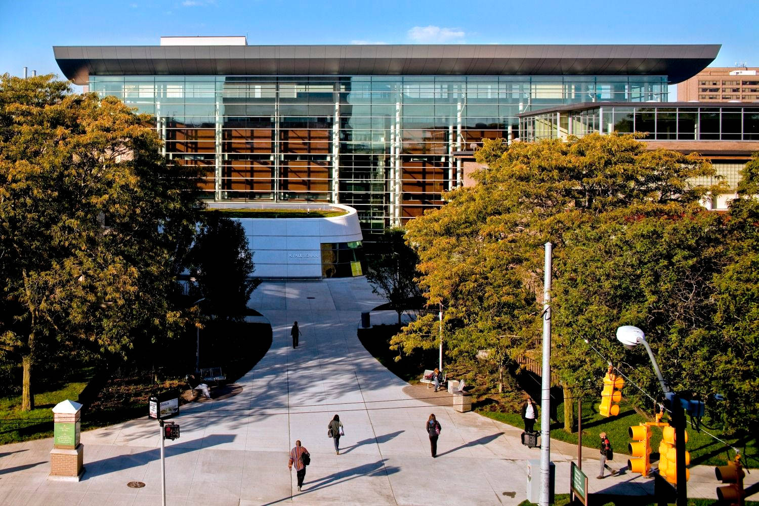 從高處俯瞰韋恩州立大學校園 壁紙 — 現代玻璃建築，弧形屋頂，周圍環繞著秋天的樹木和行人，在鋪砌的人行道上，天空清澈湛藍。
