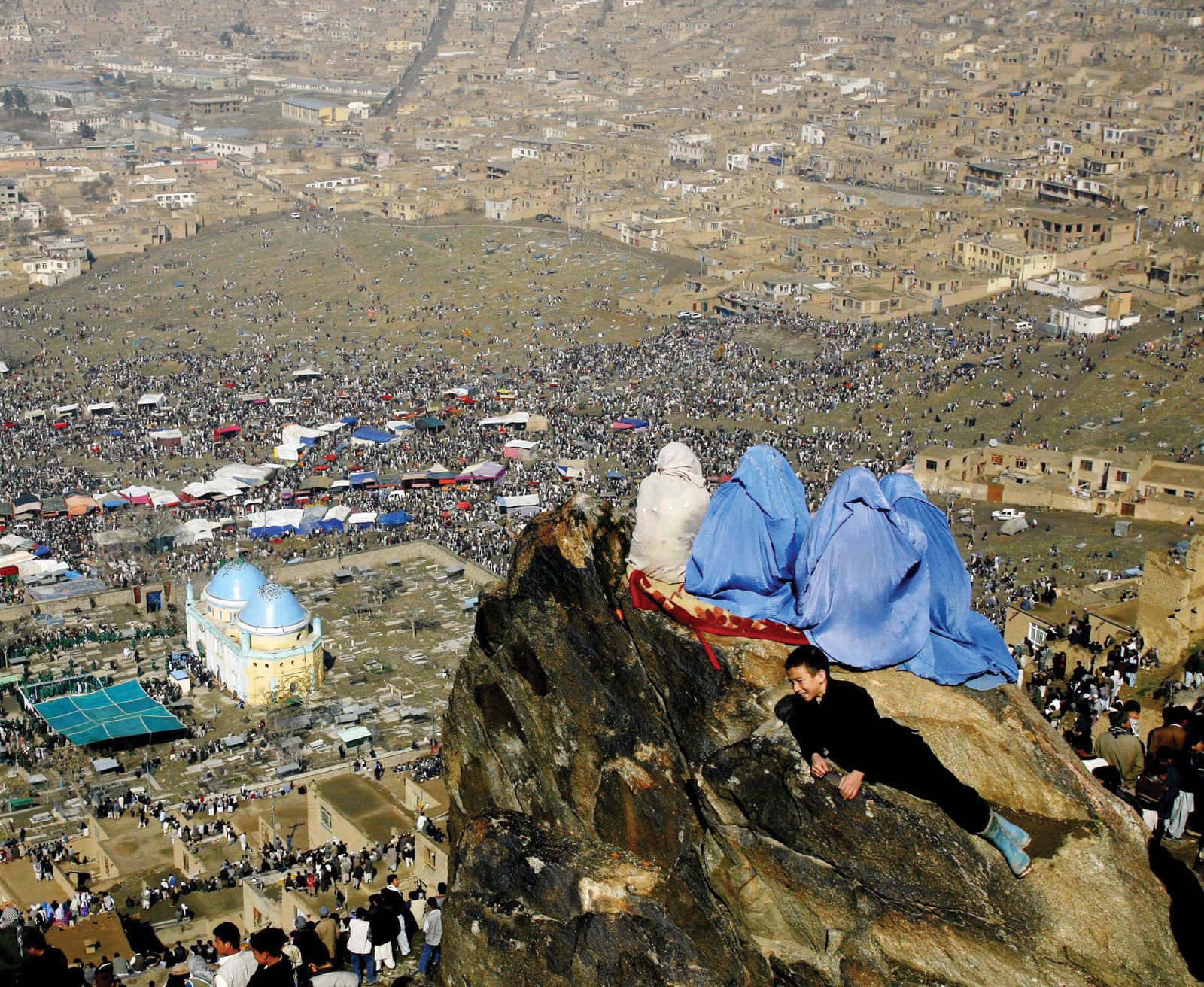Afghanistan 桌布 壁紙 — 從高角度俯瞰岩石懸崖，俯瞰陽光明媚的沙漠小鎮中聚集在藍色圓頂神社周圍的廣大人群。