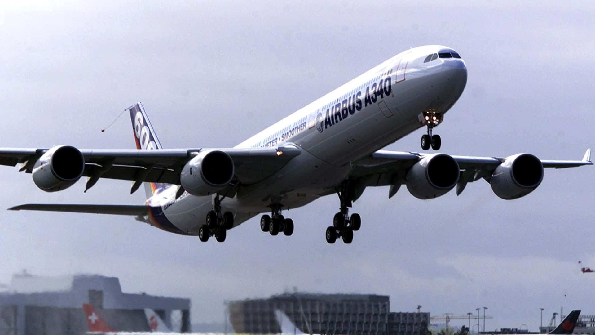 從鳥瞰視角欣賞巨型客機 壁紙 — 一架大型白色空中巴士A340飛機放下起落架，在陰天背景下接近降落，背景有城市建築物。
