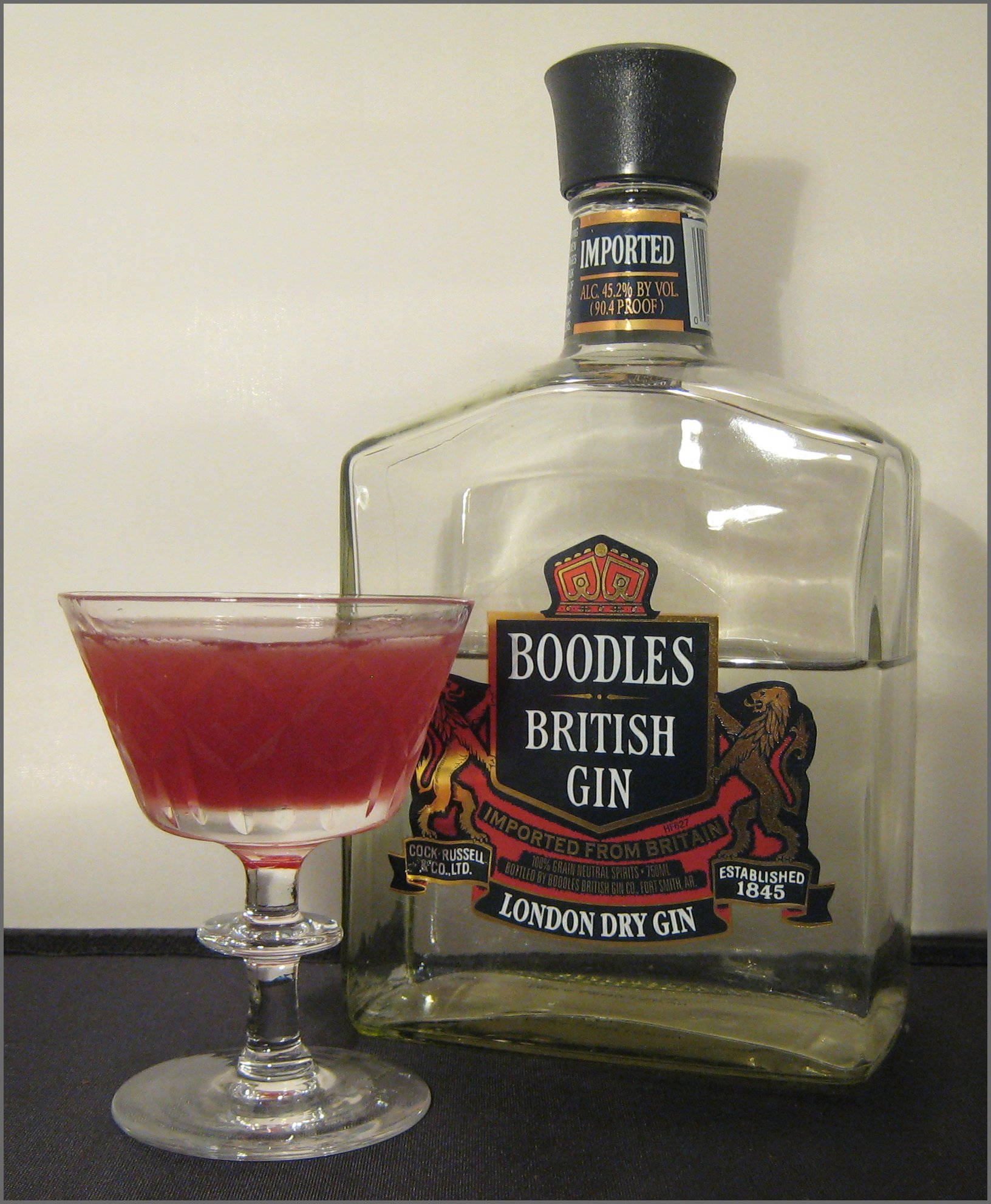 復古Boodles英國琴酒瓶 壁紙 — 一瓶透明的Boodles British Gin放在一個裝滿鮮紅色雞尾酒的碟形香檳杯旁邊，背景是純白牆壁。