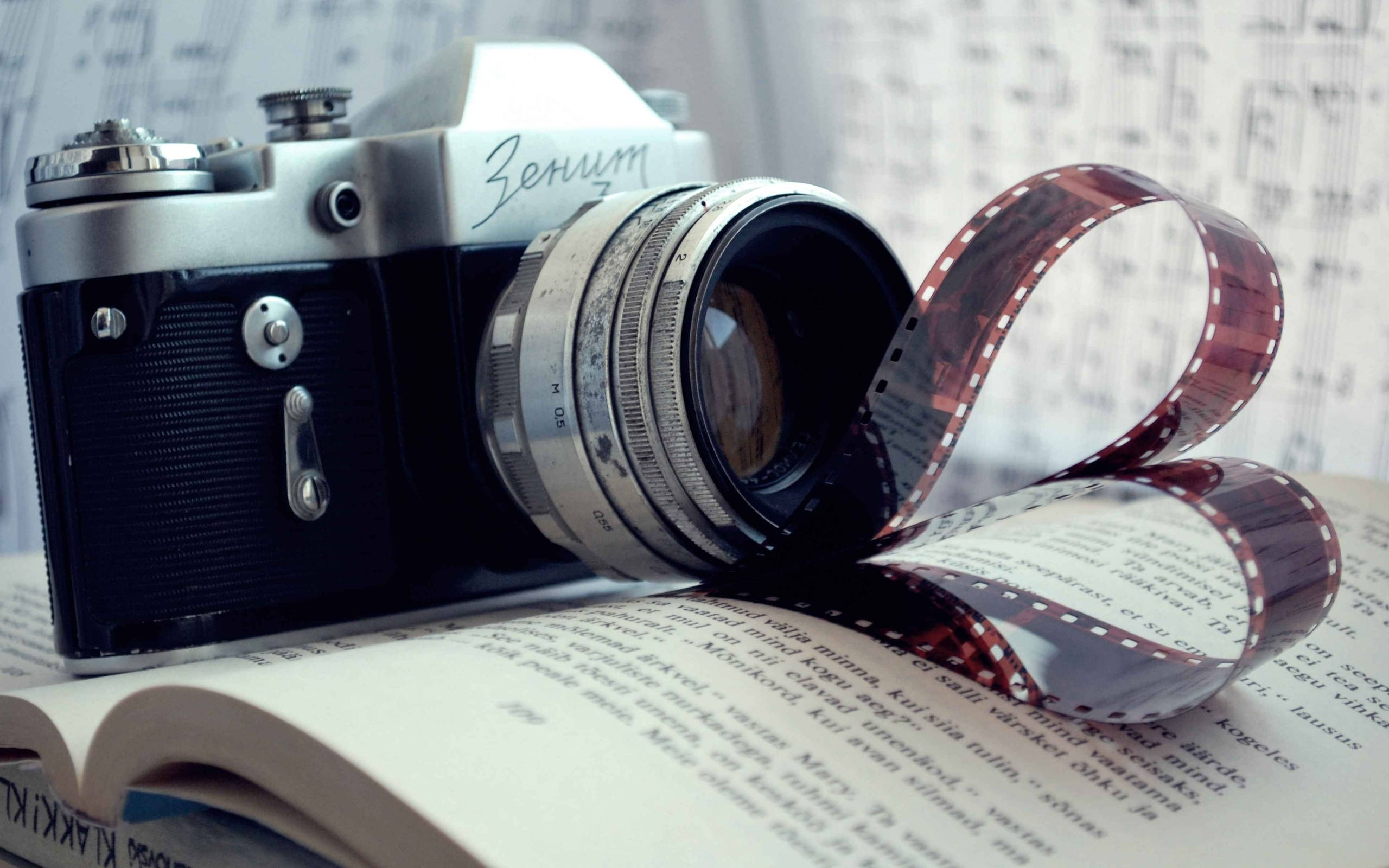 Dslr Camera 桌布 壁紙 — 復古Zenit相機擱在打開的書本上，菲林卷曲如絲帶，襯以模糊的樂譜背景。