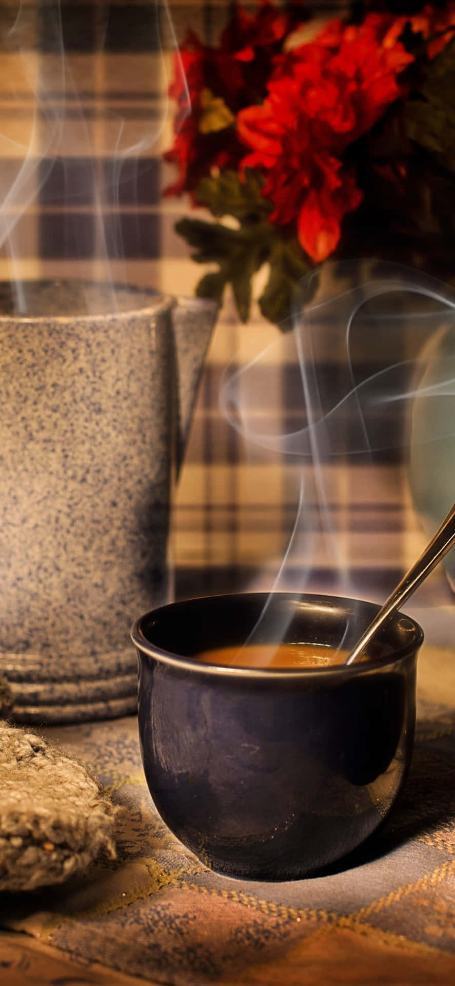 冒煙的咖啡杯和陶瓷杯放在附近帶有紅花的格紋布上。