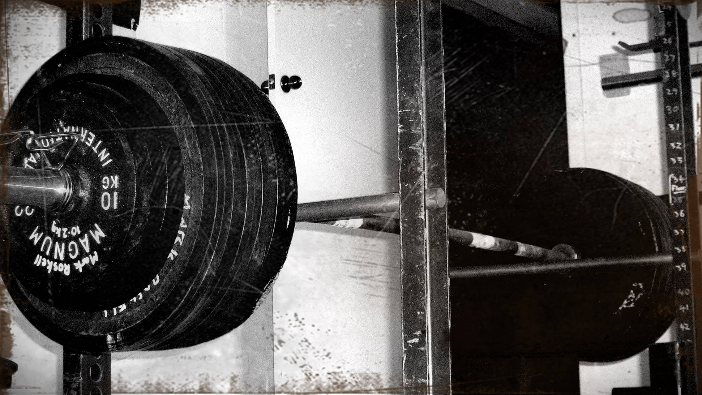 復古啞鈴放在深蹲架上 壁紙 — 一張復古黑白照片，展示一個帶有「MAGNUM」槓鈴片的沉重槓鈴，放置在磨損的健身架上。