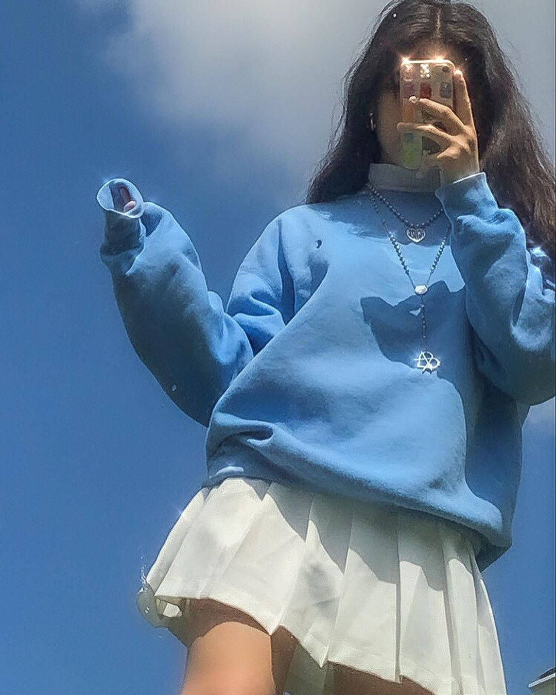 穿著藍色運動衫和白色裙子的人在戶外藍天下對著鏡子自拍。