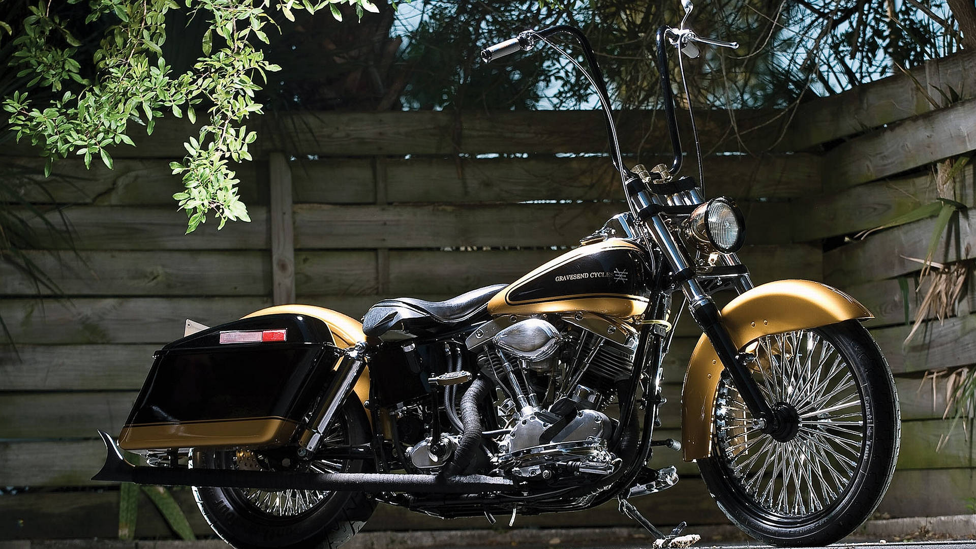 復古摩托車配哈雷戴維森標誌 壁紙 — 一架閃亮的金色和黑色Harley-Davidson電單車停泊在枝葉茂密的樹枝下，旁邊是飽經風霜的木柵欄。