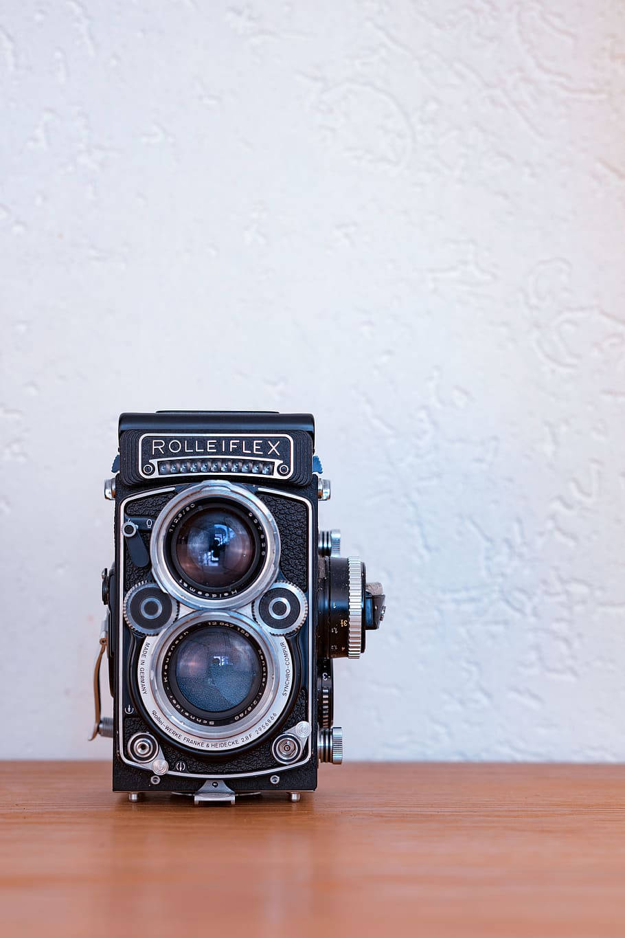 復古極簡羅利福萊克斯相機 壁紙 — 一部經典 Rolleiflex 雙鏡反光相機居中放置在木質表面上，背景是帶紋理的白色牆壁。