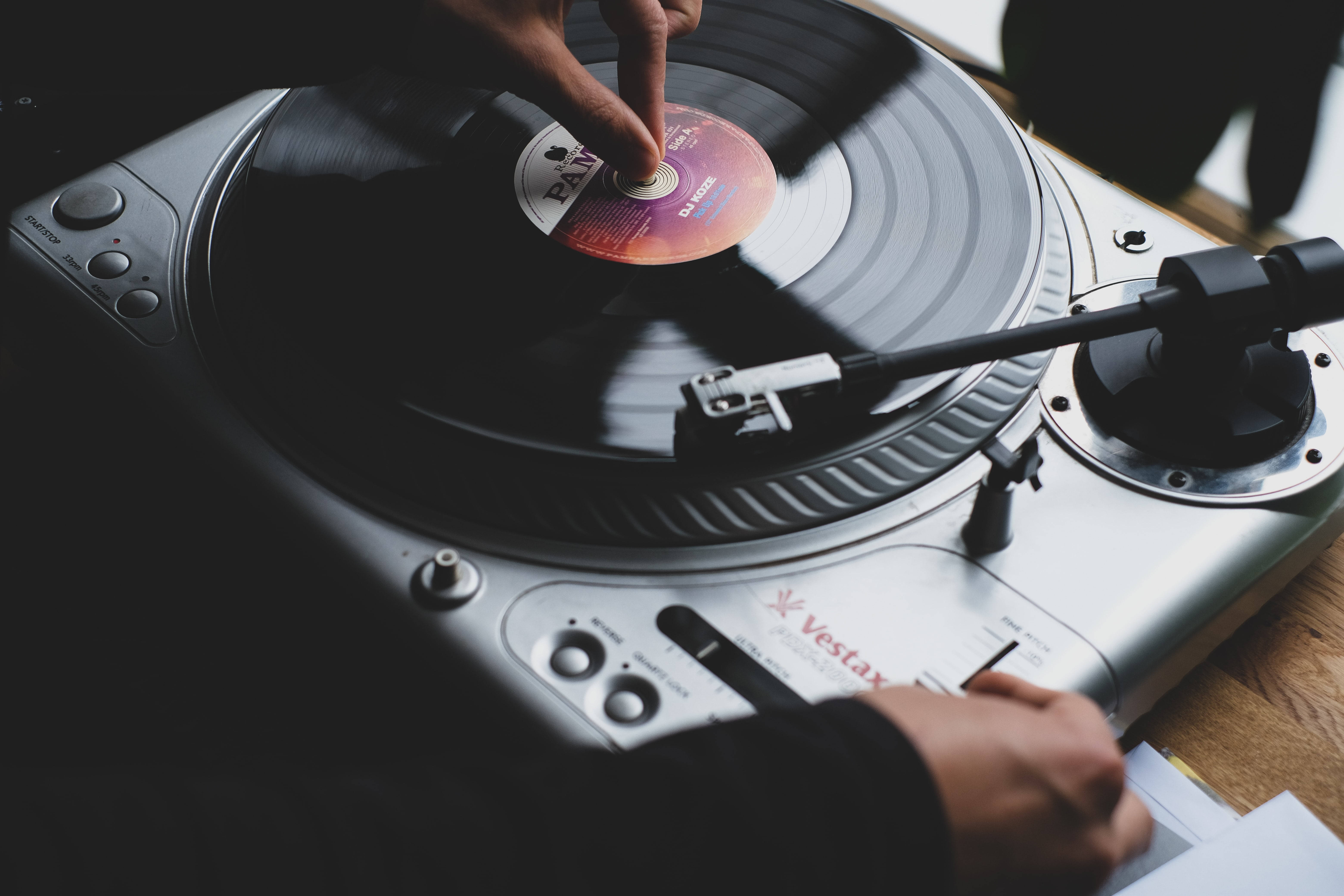 復古美學 Vestax 轉盤 壁紙 — 一隻手輕輕地將黑膠唱片放到銀色Vestax唱盤上，唱針懸浮在旋轉的唱片上方。
