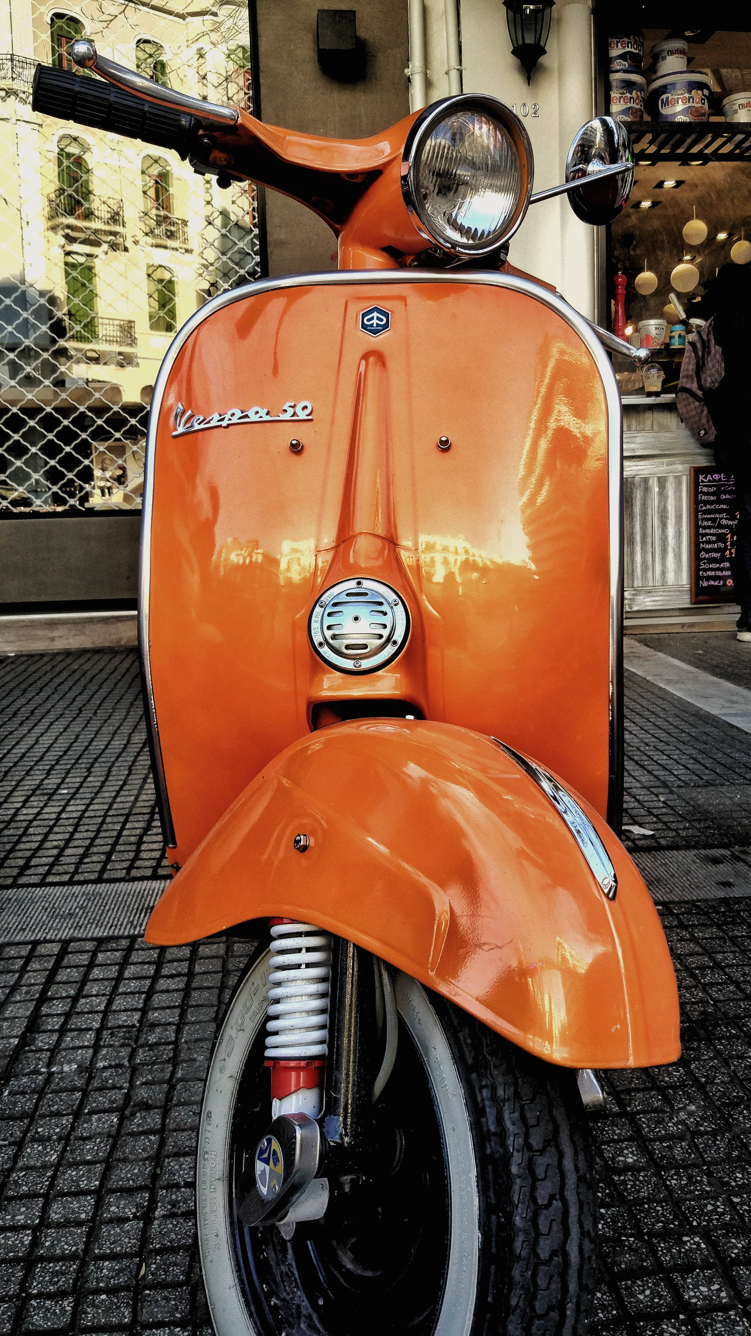 復古美學橙色維斯帕50 壁紙 — 一輛充滿活力的橙色復古Vespa 50踏板車在咖啡館旁的鵝卵石街道上，在日光下閃閃發光。