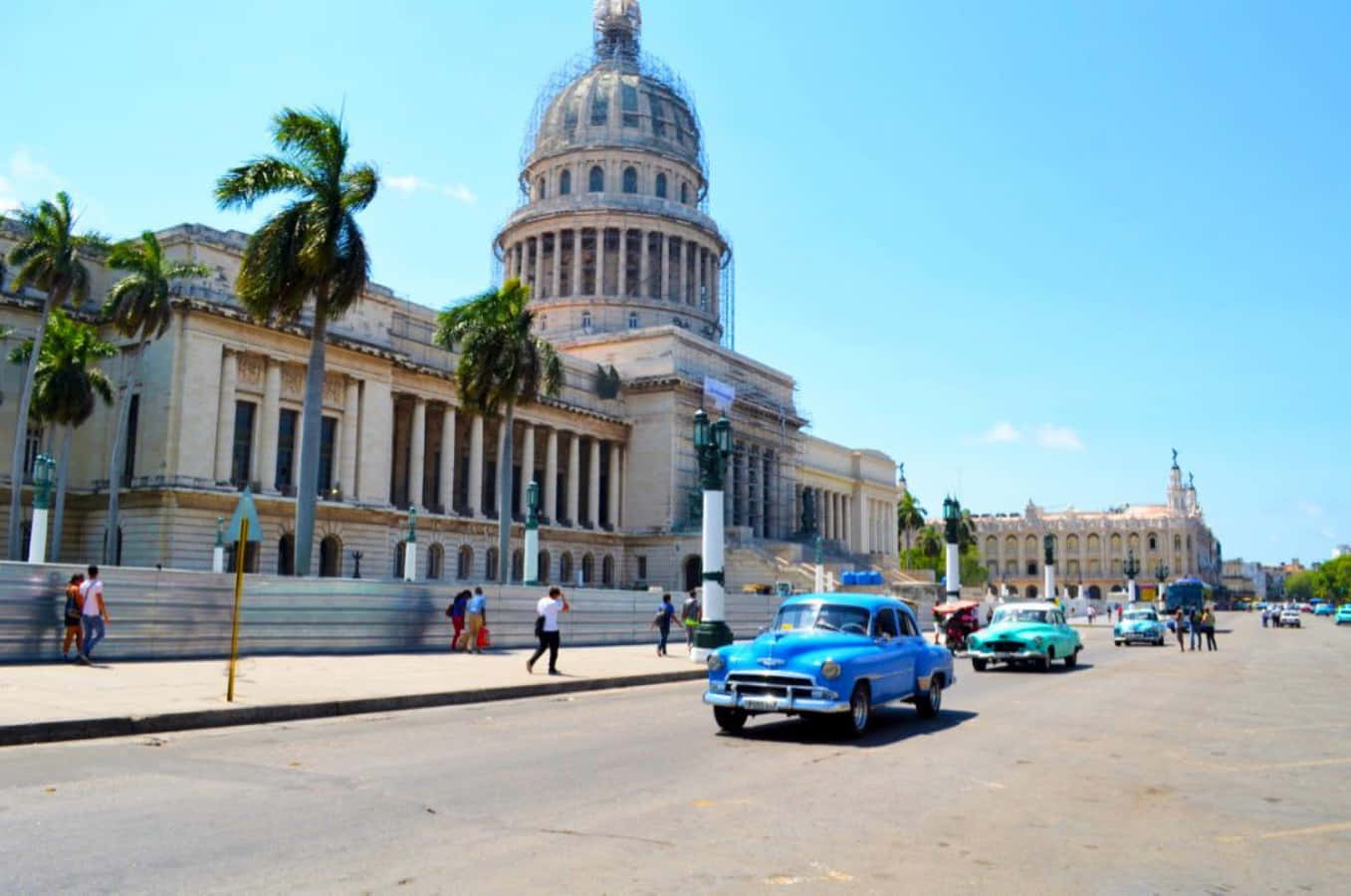 Cuba 桌布 壁紙 — 復古藍色和綠色汽車在明亮的藍天下，棕櫚樹旁駛過宏偉的新古典主義國會大廈。