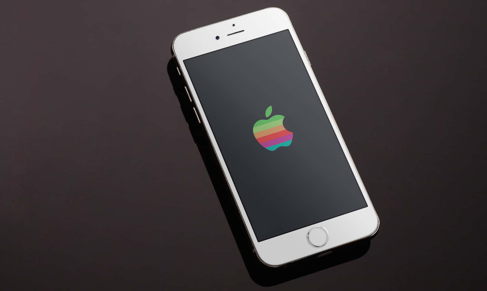 復古蘋果標誌 壁紙 — 一部白色iPhone在深色反光表面上顯示復古彩虹Apple標誌。
