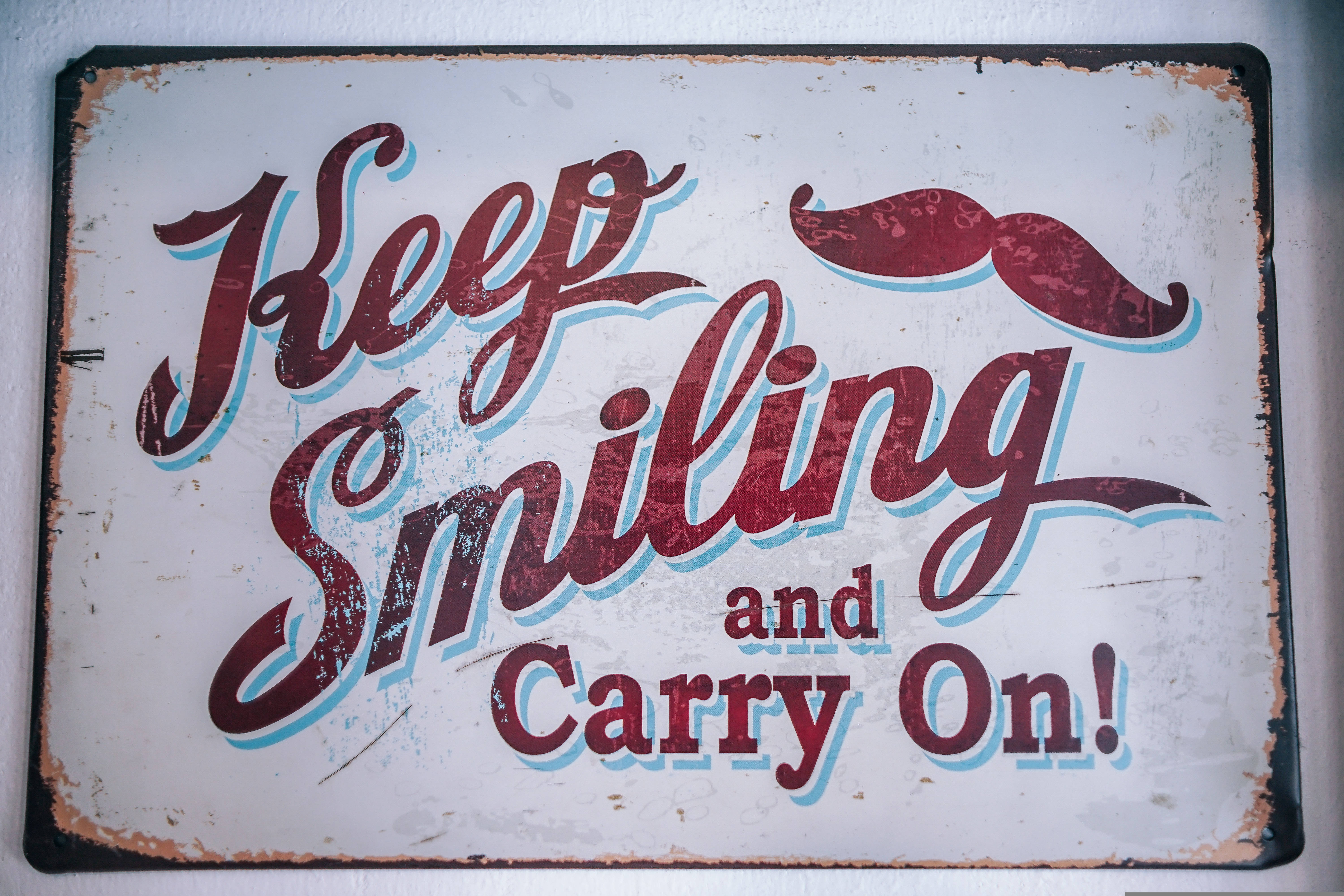 勵志語錄 桌布 壁紙 — 復古風化標誌，紅色草書文字寫著「Keep Smiling and Carry On!」，頂部有紅色鬍子。