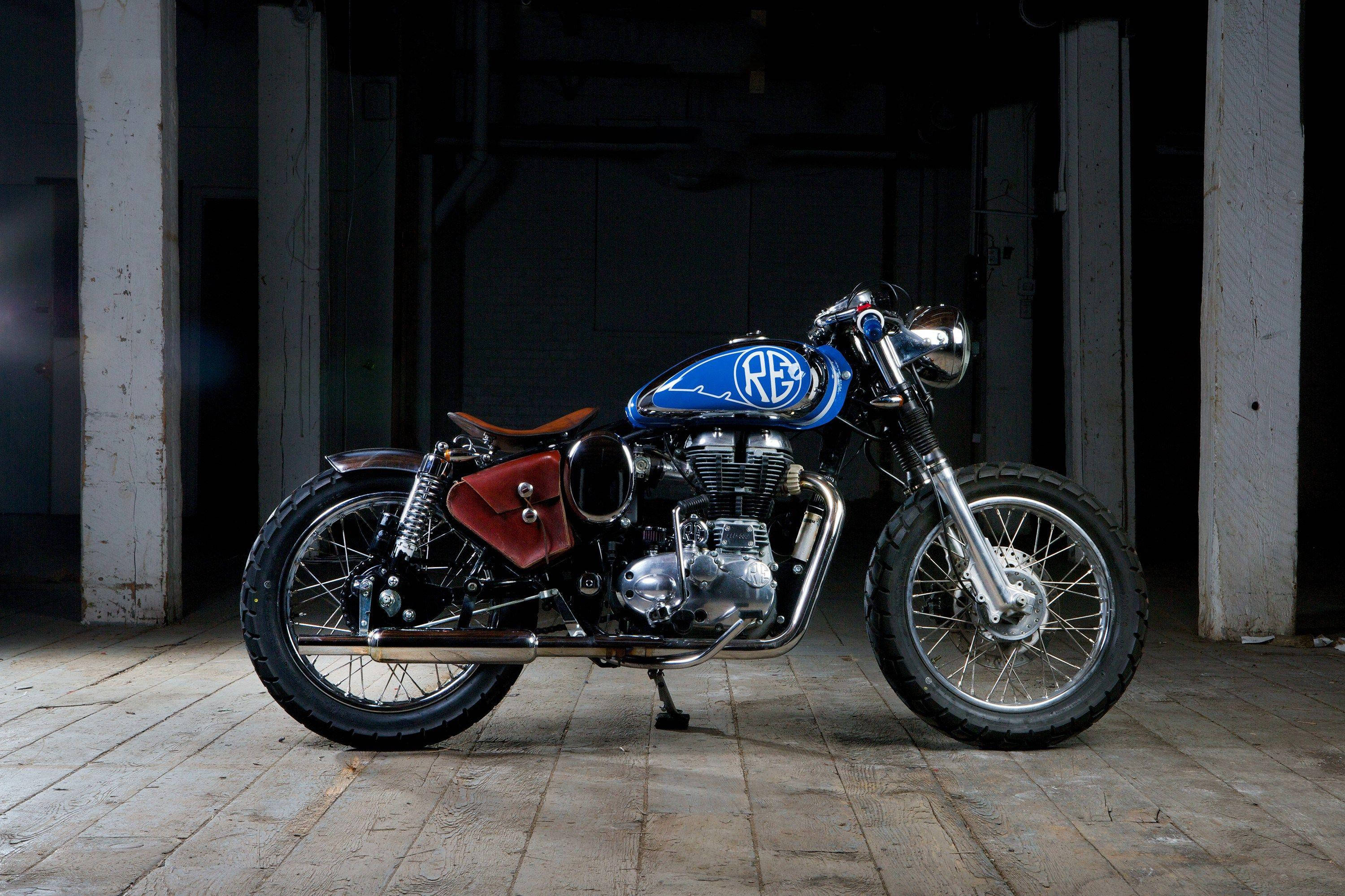 復古風情 - 海灘旁的皇家恩菲德 HD Bobber 壁紙 — 一架復古藍色電單車配紅色側袋，佇立在燈光昏暗、有混凝土地板及白色柱子的倉庫中。