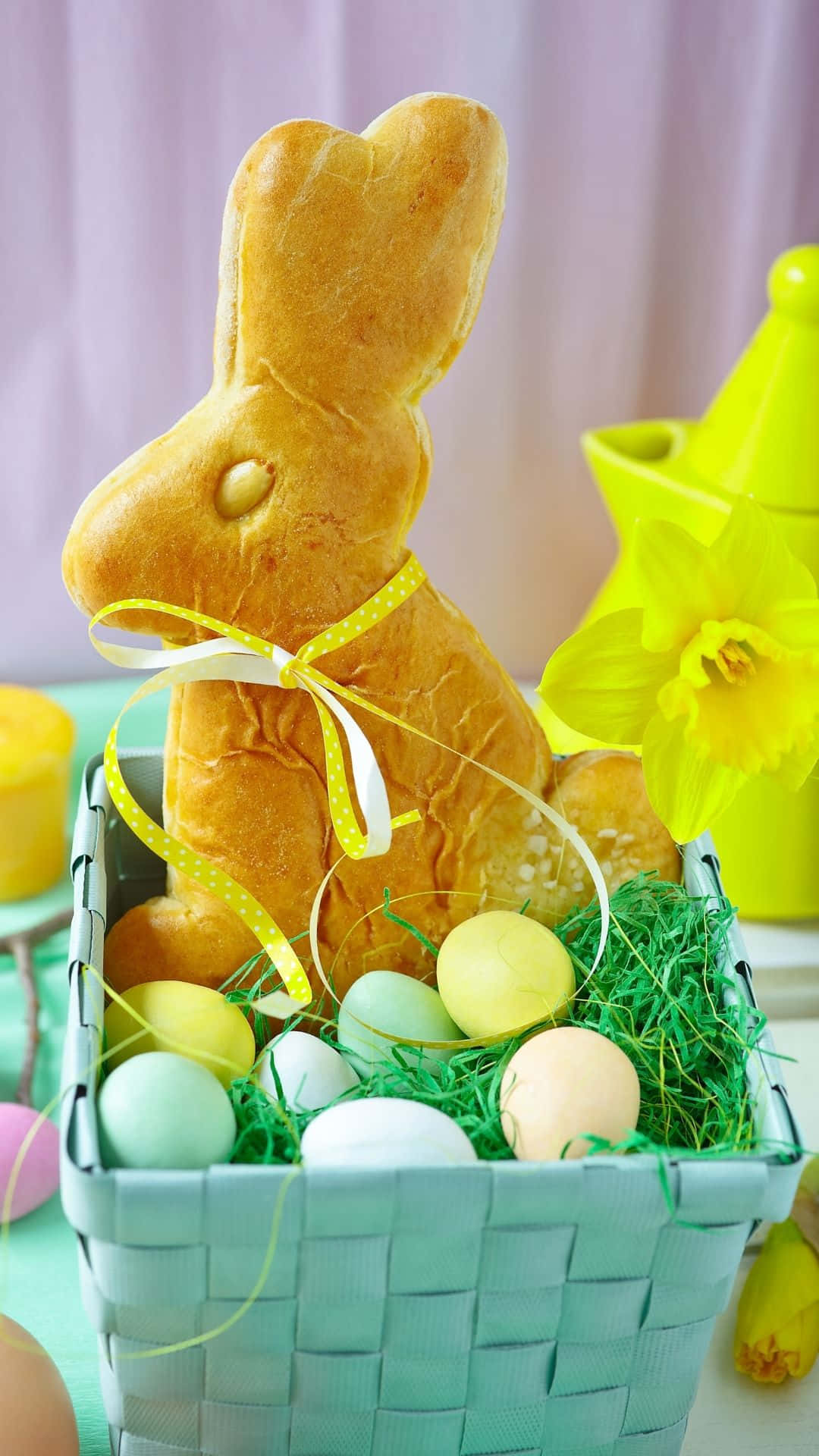 復活節兔在尋找彩蛋！ 壁紙 — 一隻金色麵包兔坐在柳條籃裡，旁邊有粉彩蛋、青草和一朵黃色水仙花。