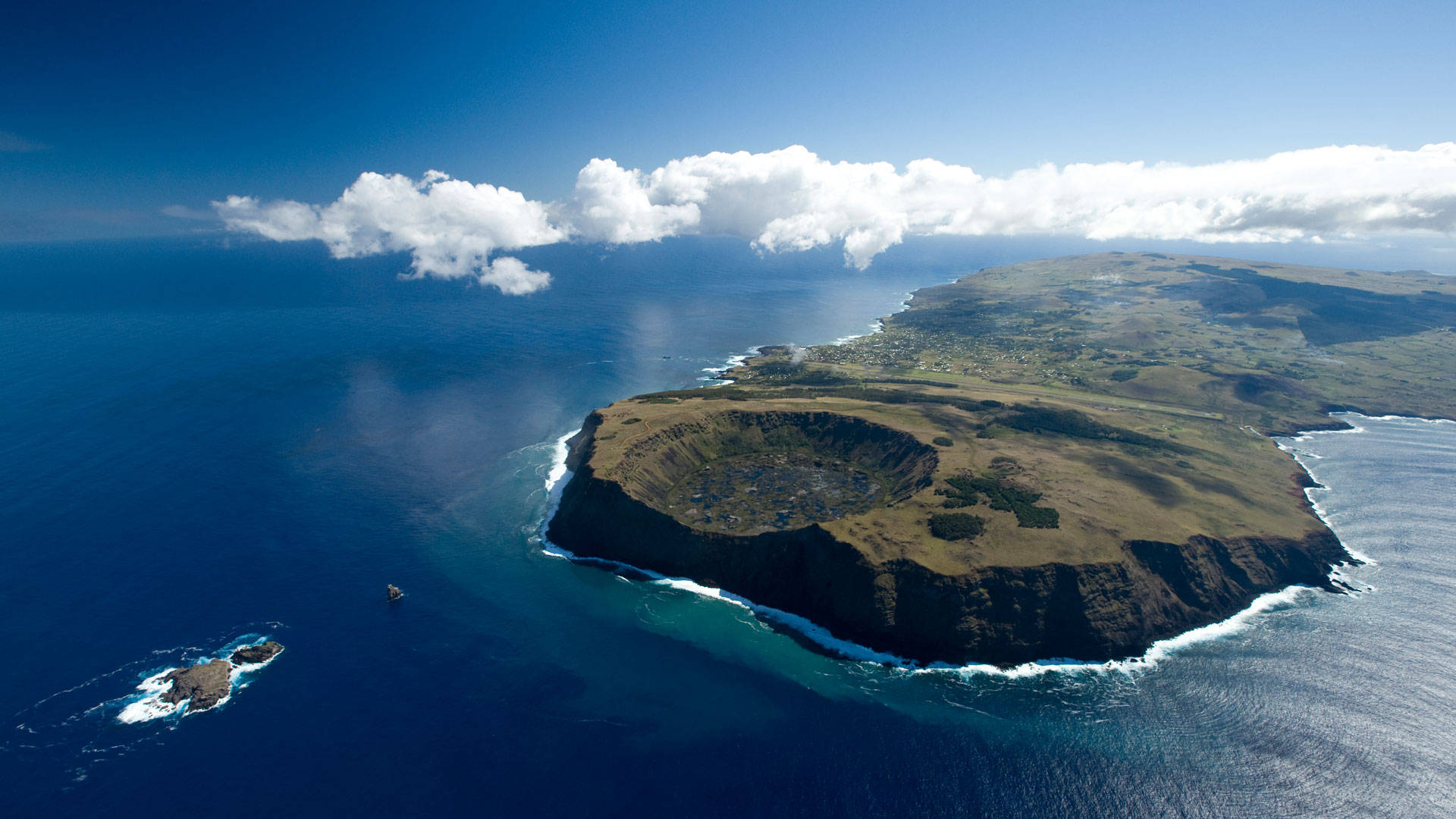 復活節島摩艾石像壯觀景觀 壁紙 — 火山口島嶼的鳥瞰圖，擁有陡峭的懸崖，被深藍色海洋和白色泡沫波浪環繞，天空明亮，雲朵散佈。