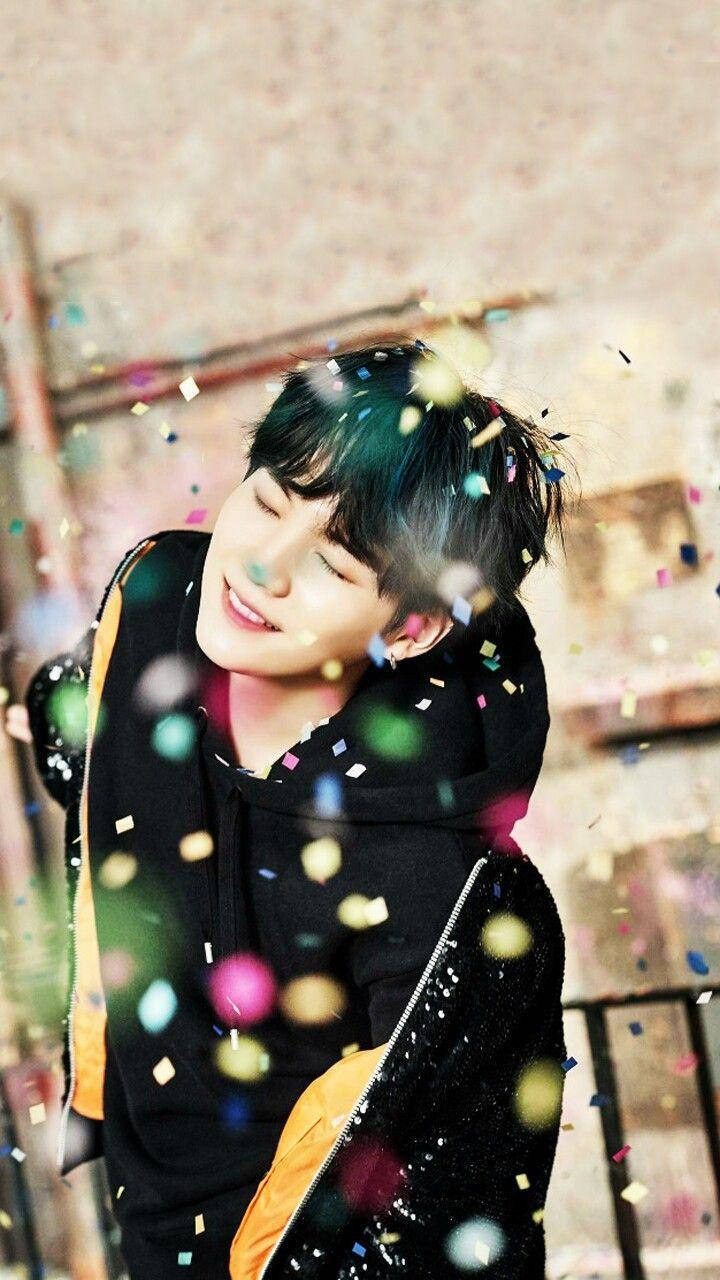 微笑 Suga BTS 壁紙 — 一名年輕男子閉上眼睛，在五彩紙屑如雨般灑落時開心地微笑。
