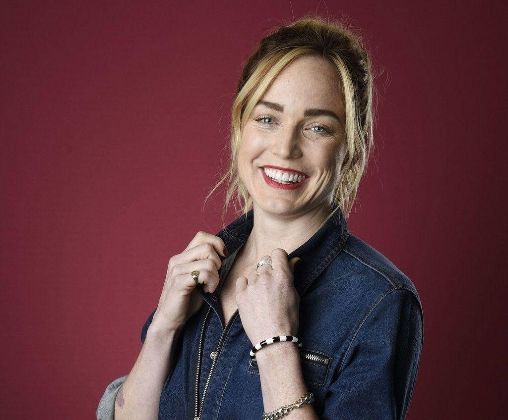 微笑的 Caity Lotz 壁紙 — 一位微笑的金髮紅唇女性，身穿牛仔外套，在深酒紅色背景前擺姿勢。