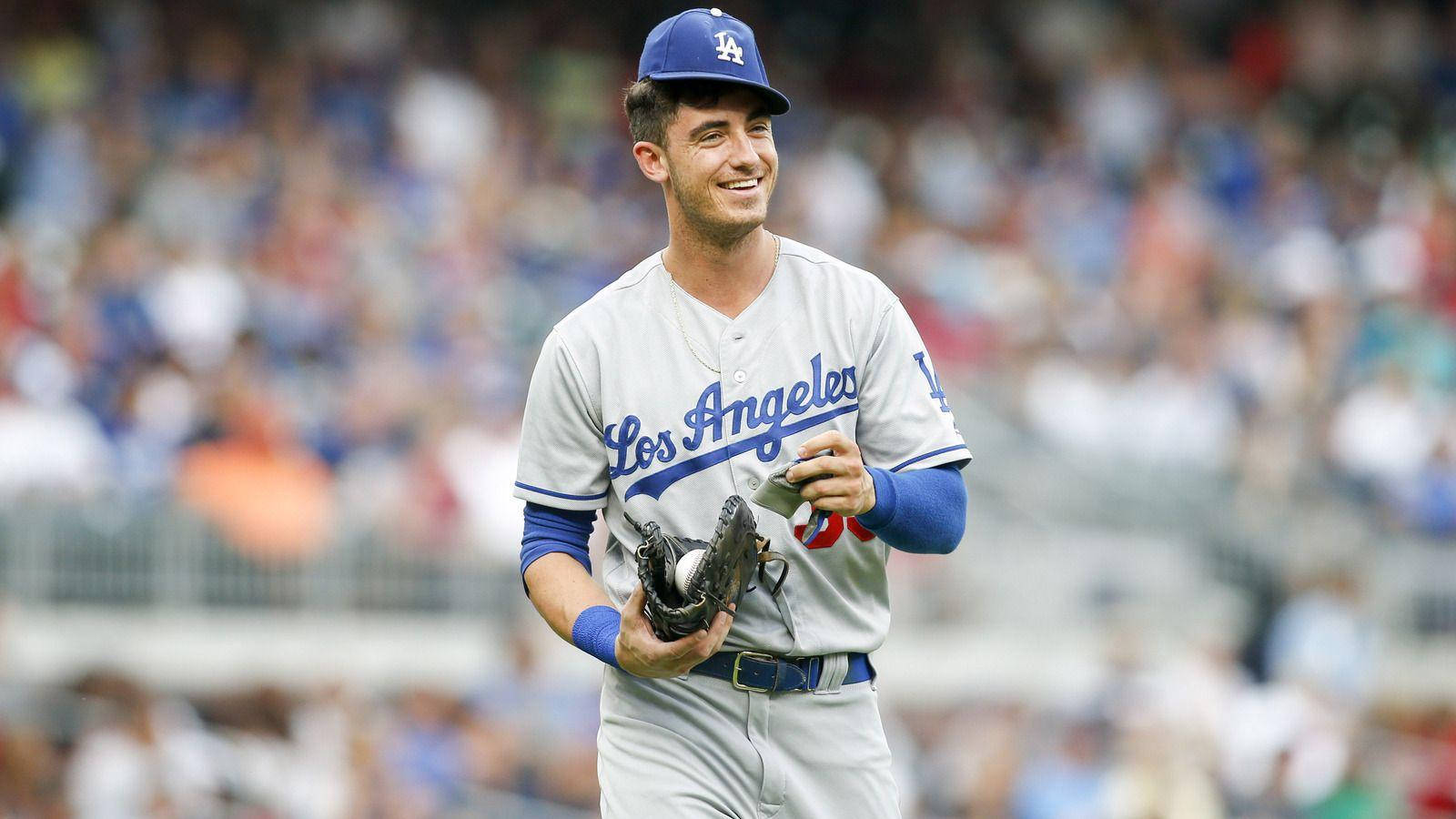 微笑的 Cody Bellinger 壁紙 — 一位微笑的洛杉磯道奇隊棒球運動員，身穿制服，手持手套和球，在陽光明媚的球場上行走，背景是模糊的球迷。