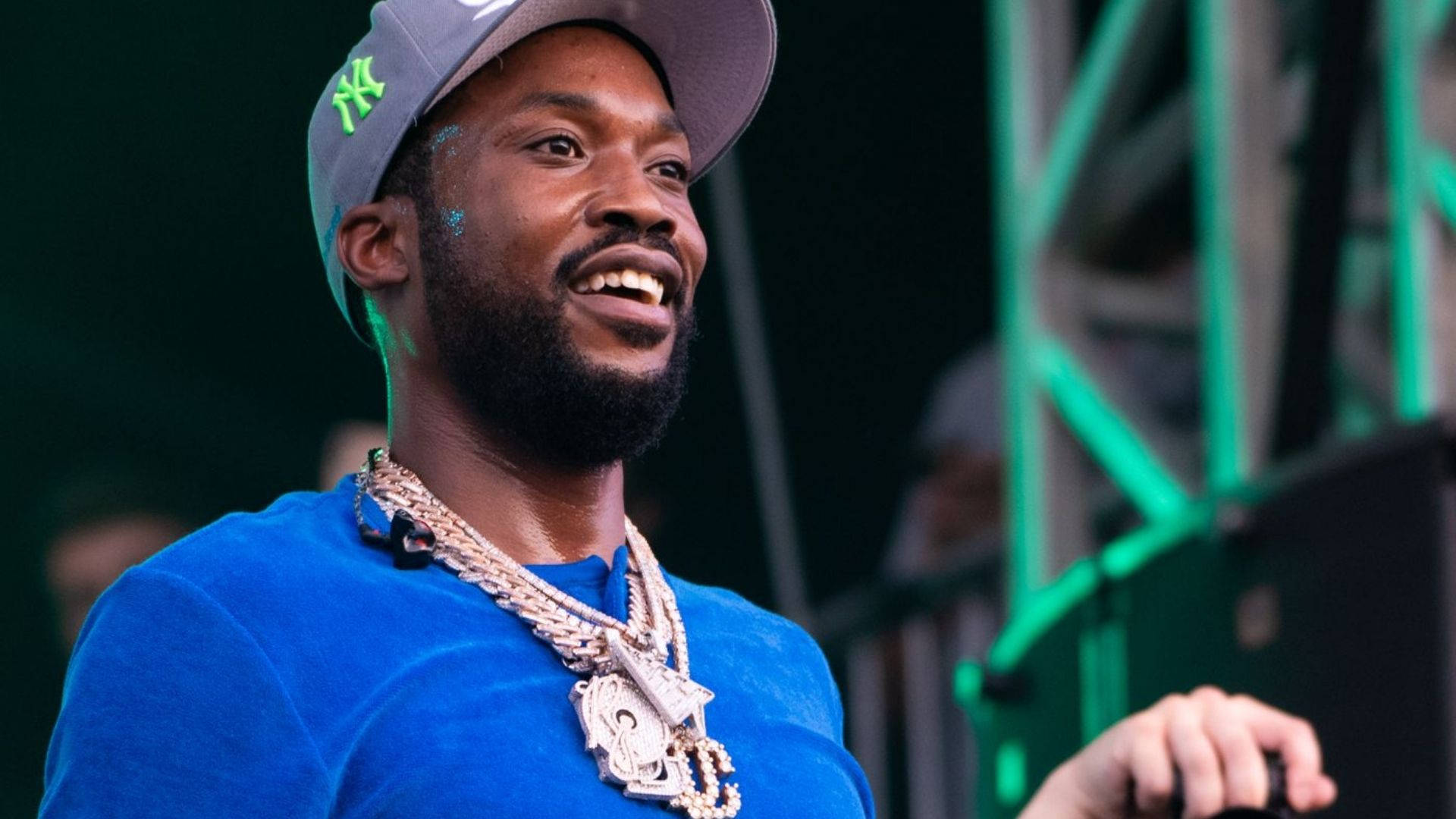 微笑的Meek Mill 壁紙 — 一個微笑的男人穿著藍色襯衫和NY帽子，在綠色舞台燈光下戴著閃亮的金鏈。