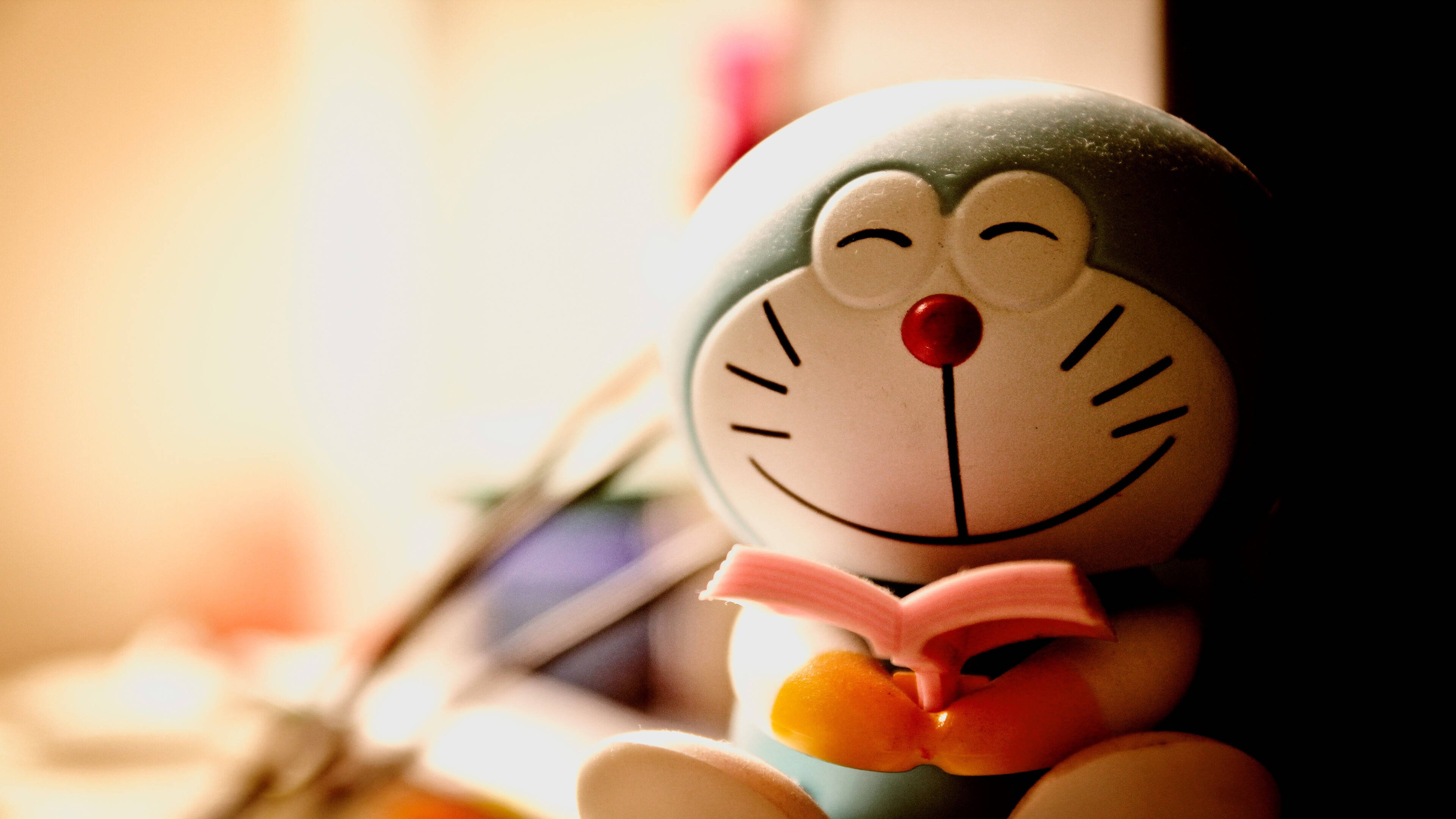 Doraemon 桌布 壁紙 — 微笑的哆啦A夢公仔在溫暖背光及柔和散景背景下拿著一本粉紅色的書。