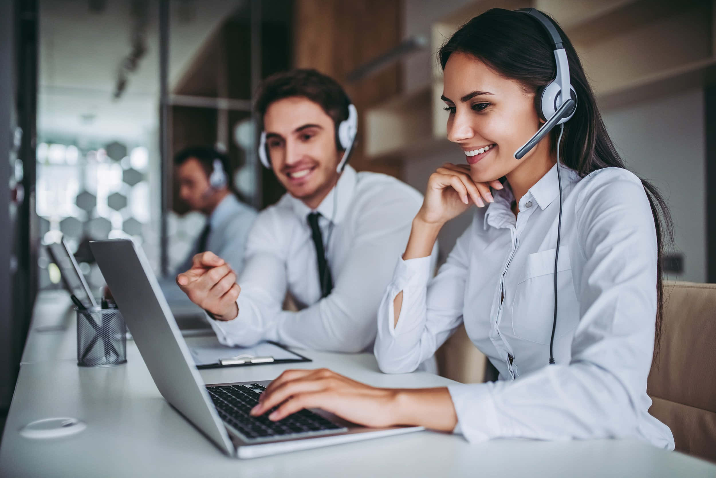 Telemarketing 桌布 壁紙 — 微笑的客戶服務人員戴著耳機，在設有玻璃隔斷和溫暖照明的現代辦公室中使用筆記本電腦工作。