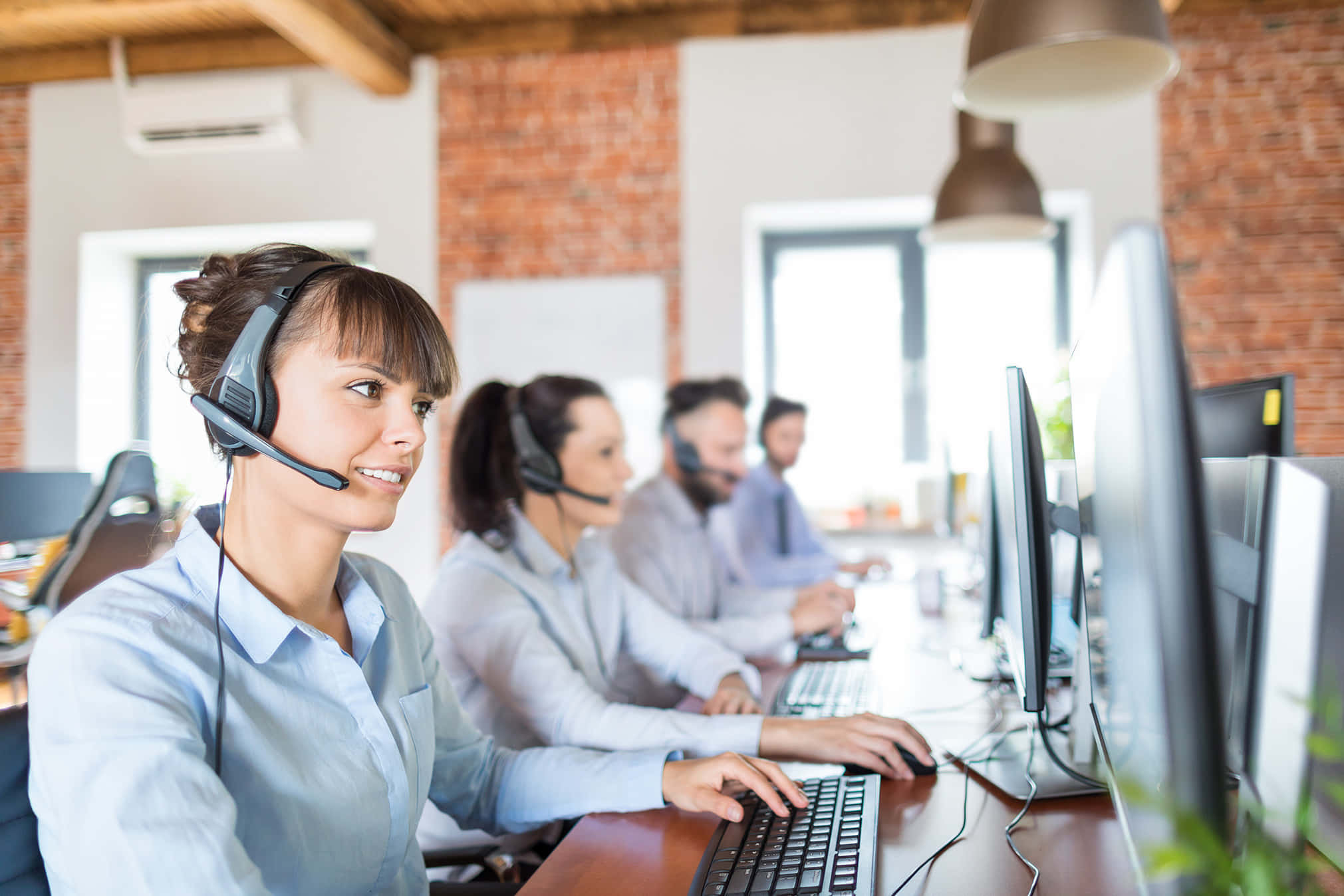Telemarketing 桌布 壁紙 — 微笑的客戶服務代理戴著耳機，在設有磚牆和同事的現代開放式辦公室中使用電腦工作。