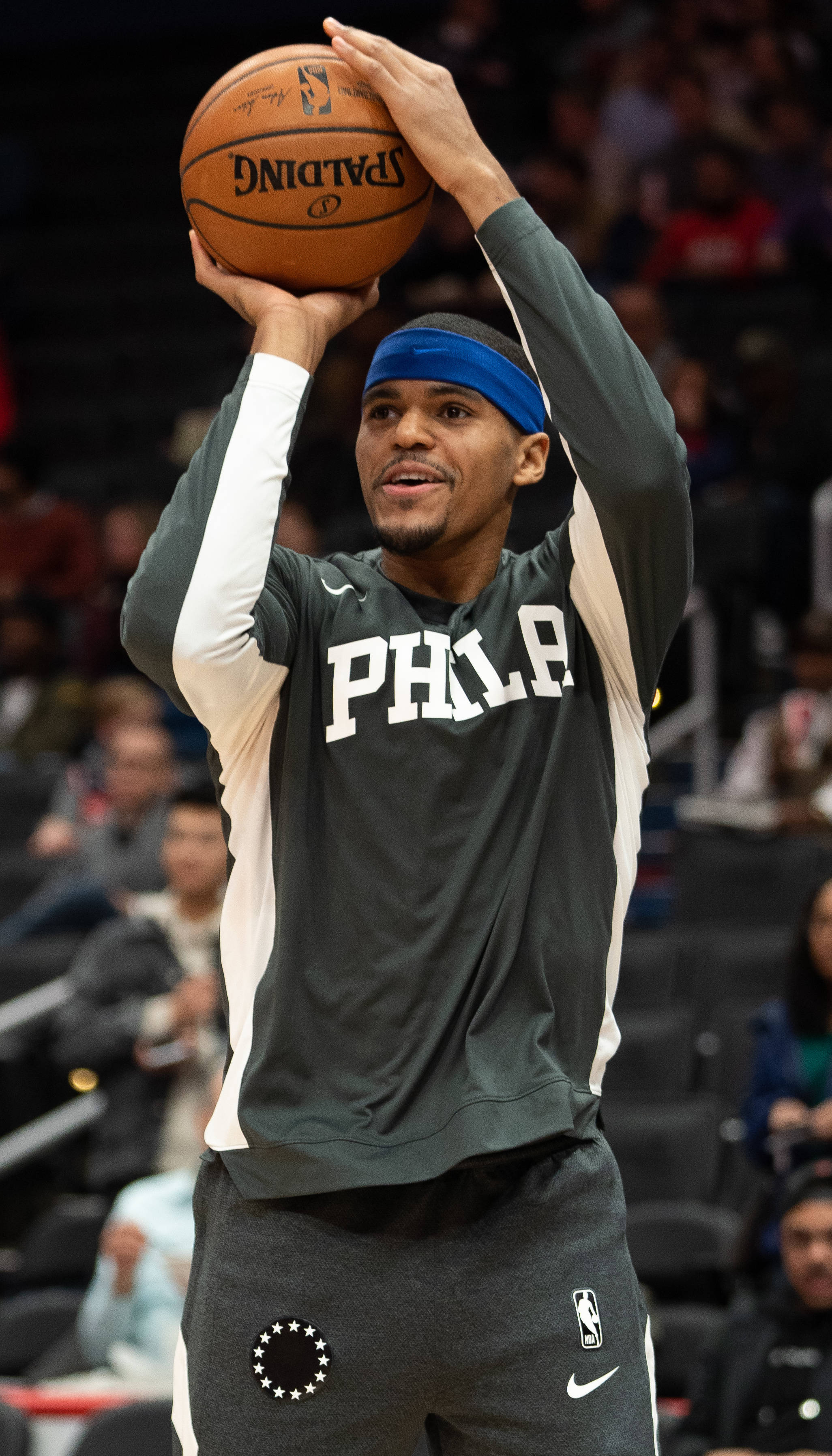 微笑的托拜厄斯·哈里斯NBA球員費城76人隊 壁紙 — 一名身穿費城球衣的籃球運動員在比賽中準備投擲Spalding籃球，背景是模糊的觀眾。