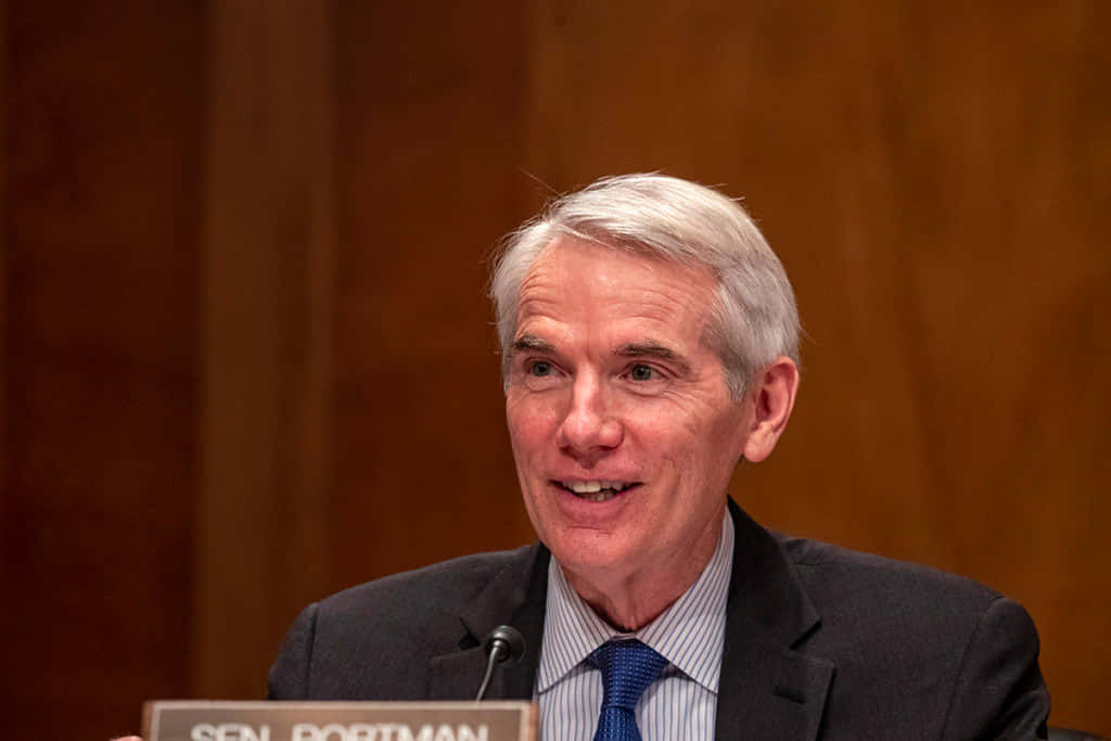 微笑的羅布·波特曼 壁紙 — 參議員 Rob Portman 在正式聽證會上微笑發言，背景為木質牆面。