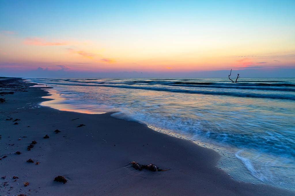 德克薩斯州帕德雷島國家海灘 壁紙 — 夕陽將天空染成柔和的橙色和粉紅色調，溫柔的海浪拍打著沙灘，沙灘上散落著漂流木。