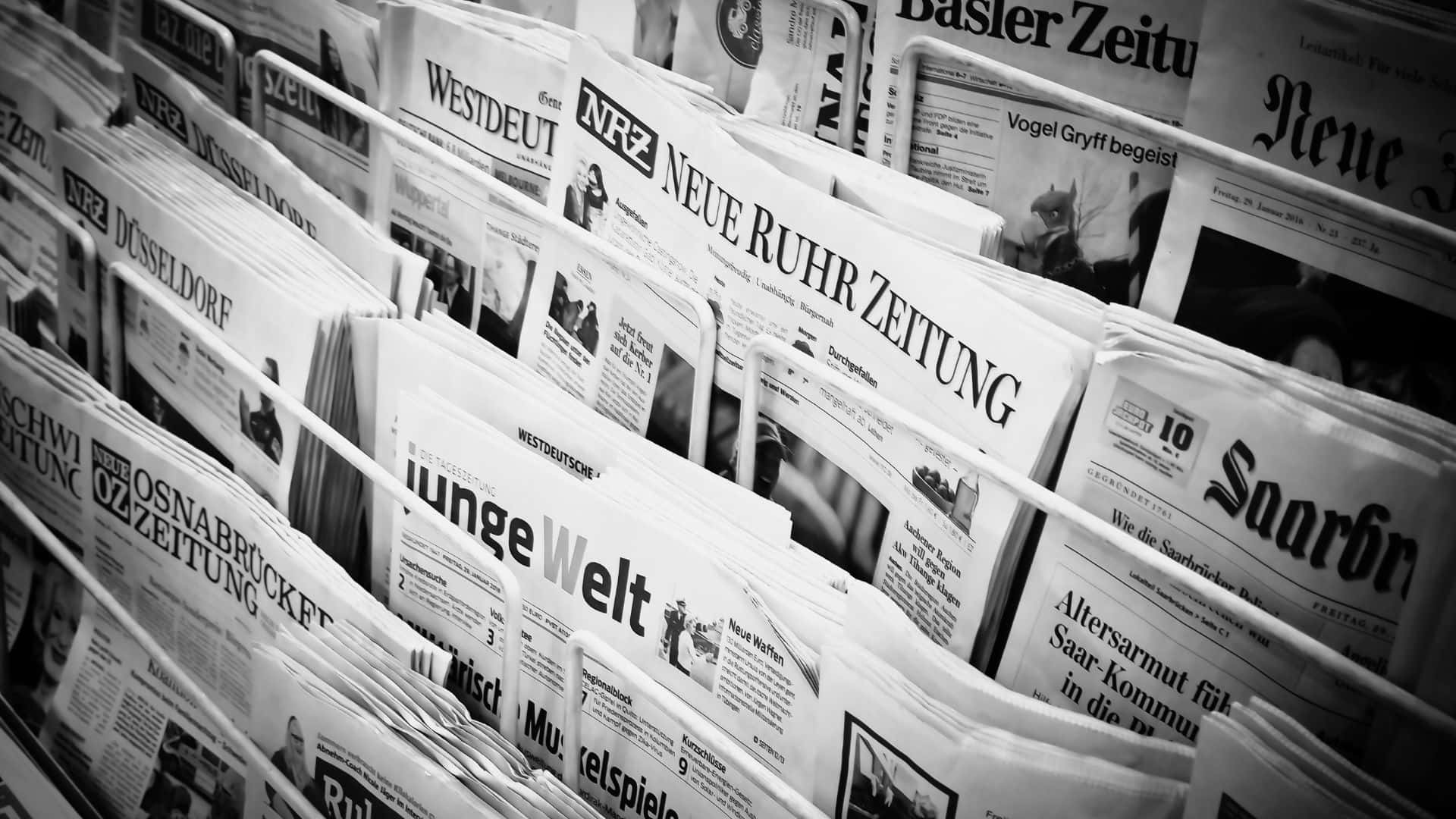 Black And White Newspaper 桌布 壁紙 — 德國報紙堆疊在報架上的黑白特寫，可見標題和文字。