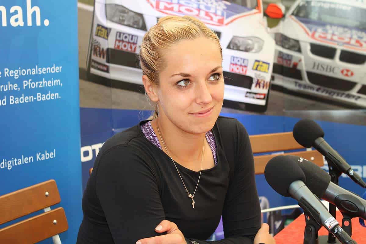 德國網球選手Sabine Lisicki訪問照 壁紙 — 一位金髮女子身穿黑色上衣，坐在記者會上微笑，身後有賽車海報。