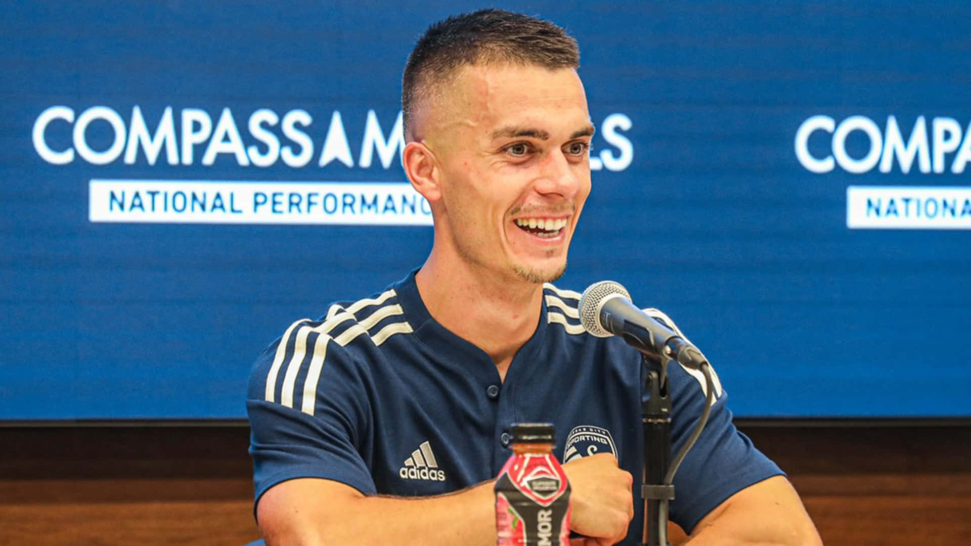 德國職業足球員埃里克·托米 壁紙 — 一位微笑的男子身穿深藍色Adidas Polo衫，在藍色「Compass Minerals」背景的新聞發布會上手持紅色運動飲料瓶。