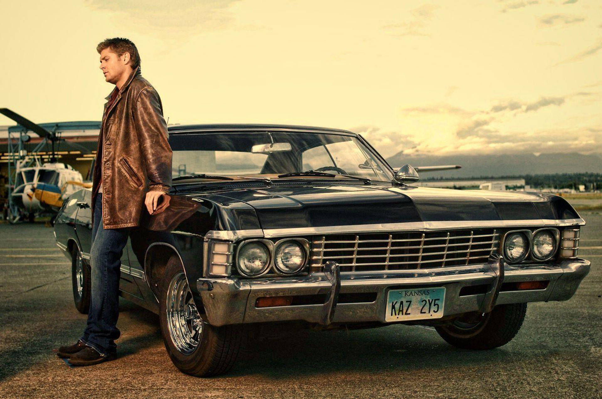 德恩·溫徹斯特的 1967 年雪佛蘭 Impala 壁紙 — 一名身穿棕色皮褸的男子在黃金時段的機場停機坪上，靠在一輛帶有鍍鉻細節的黑色經典汽車上。