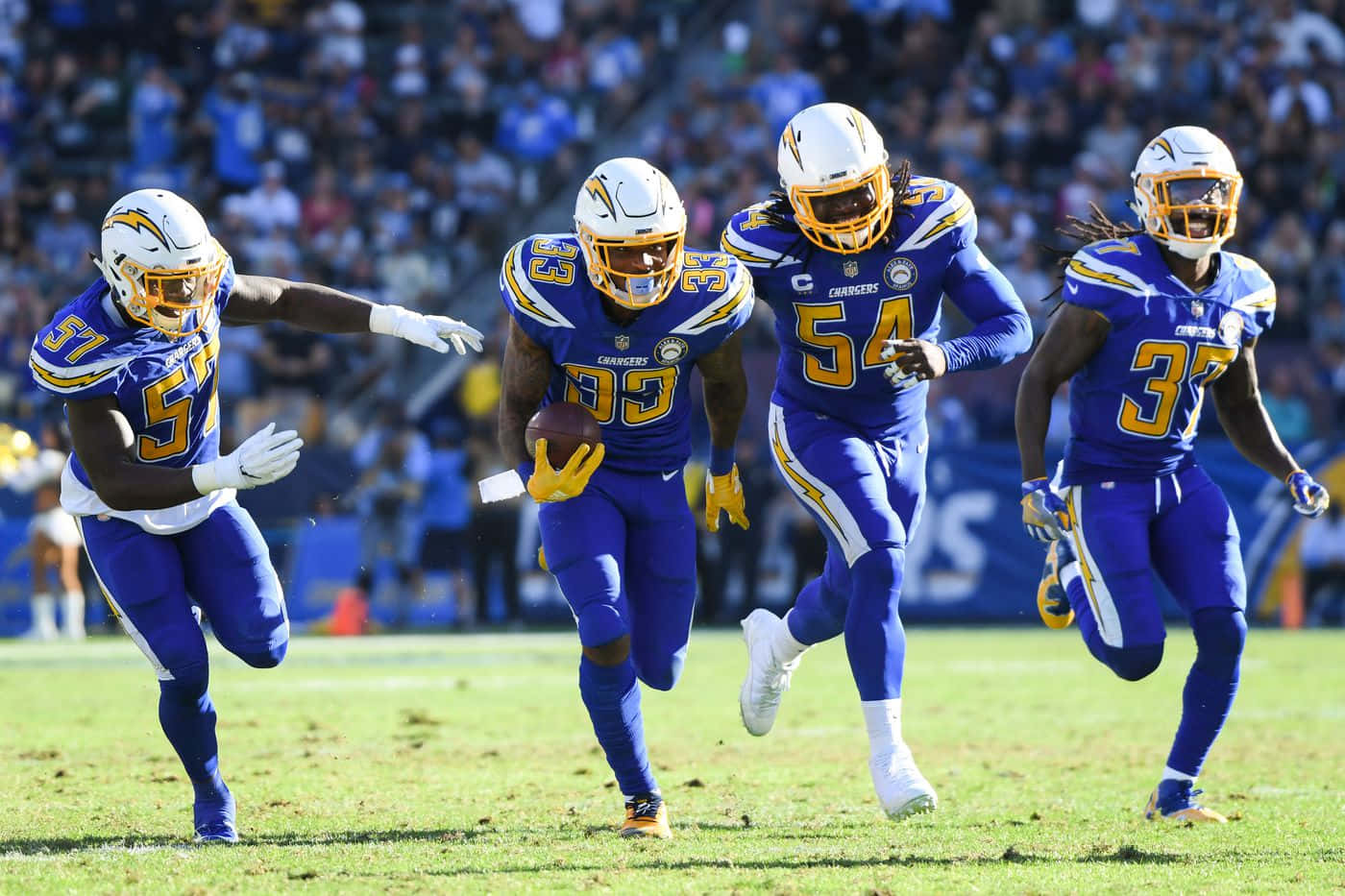 德文·詹姆斯與隊友 洛杉磯chargers 壁紙 — 四名身穿藍色和金色球衣的 Chargers 美式足球員在陽光明媚的球場上全力衝刺，其中一人持球。